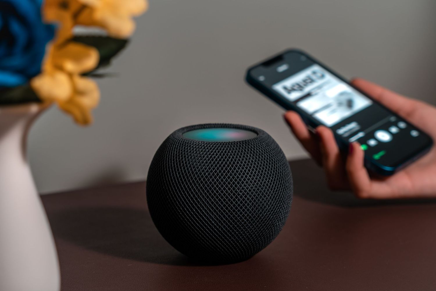 Person interacting with HomePod Mini while using smartphone in dimly lit setting.