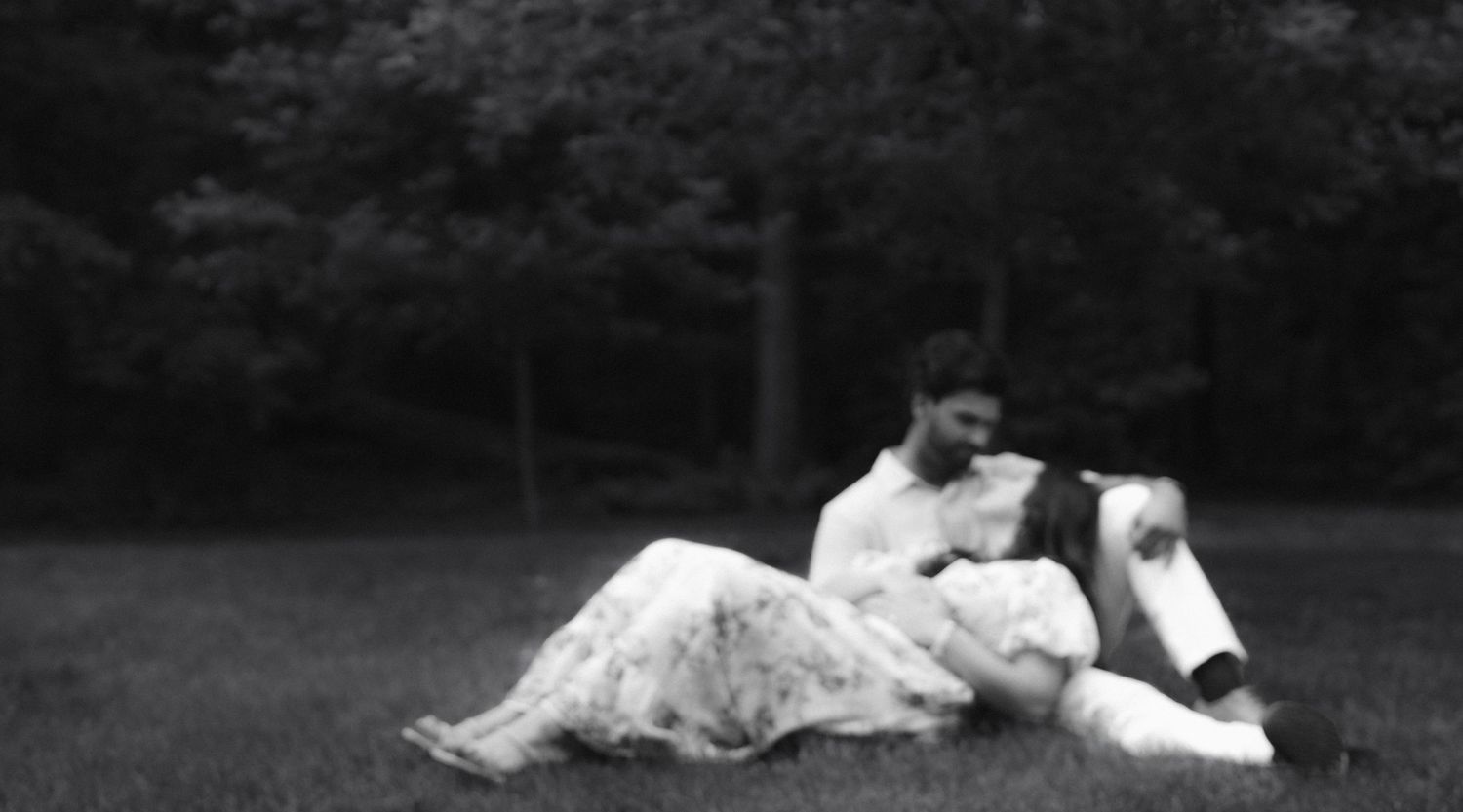 Black and white photo of a couple relaxing on grass in a park, woman in floral dress leaning against man.
