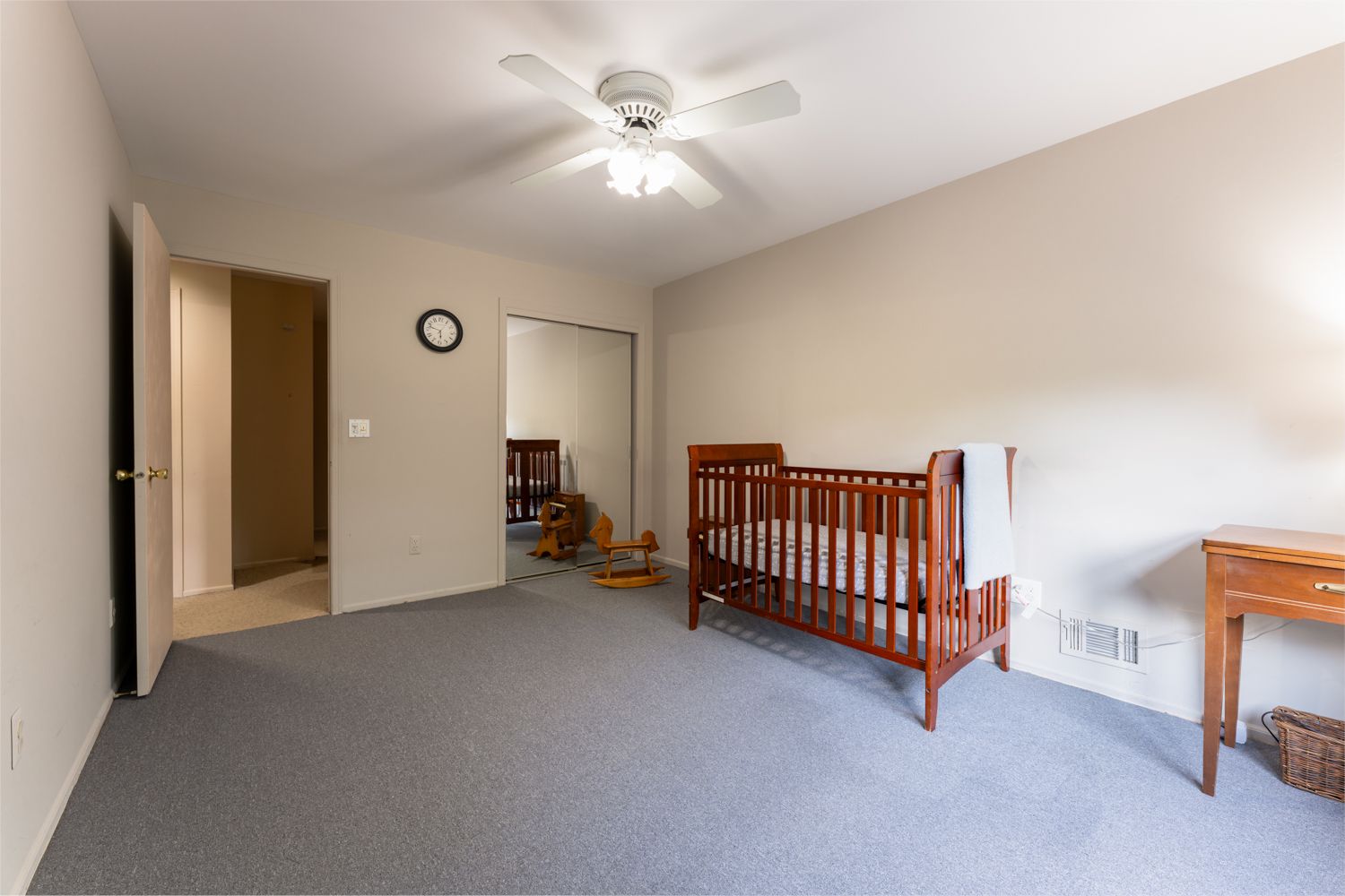 Spacious bedroom with wooden baby crib, gray carpet, ceiling fan, and side table against beige walls.
