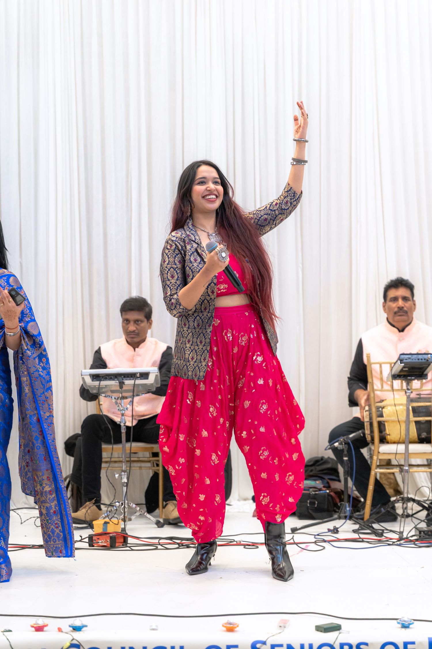 A performer in a bright red traditional outfit dances energetically on stage with musicians visible in the background.