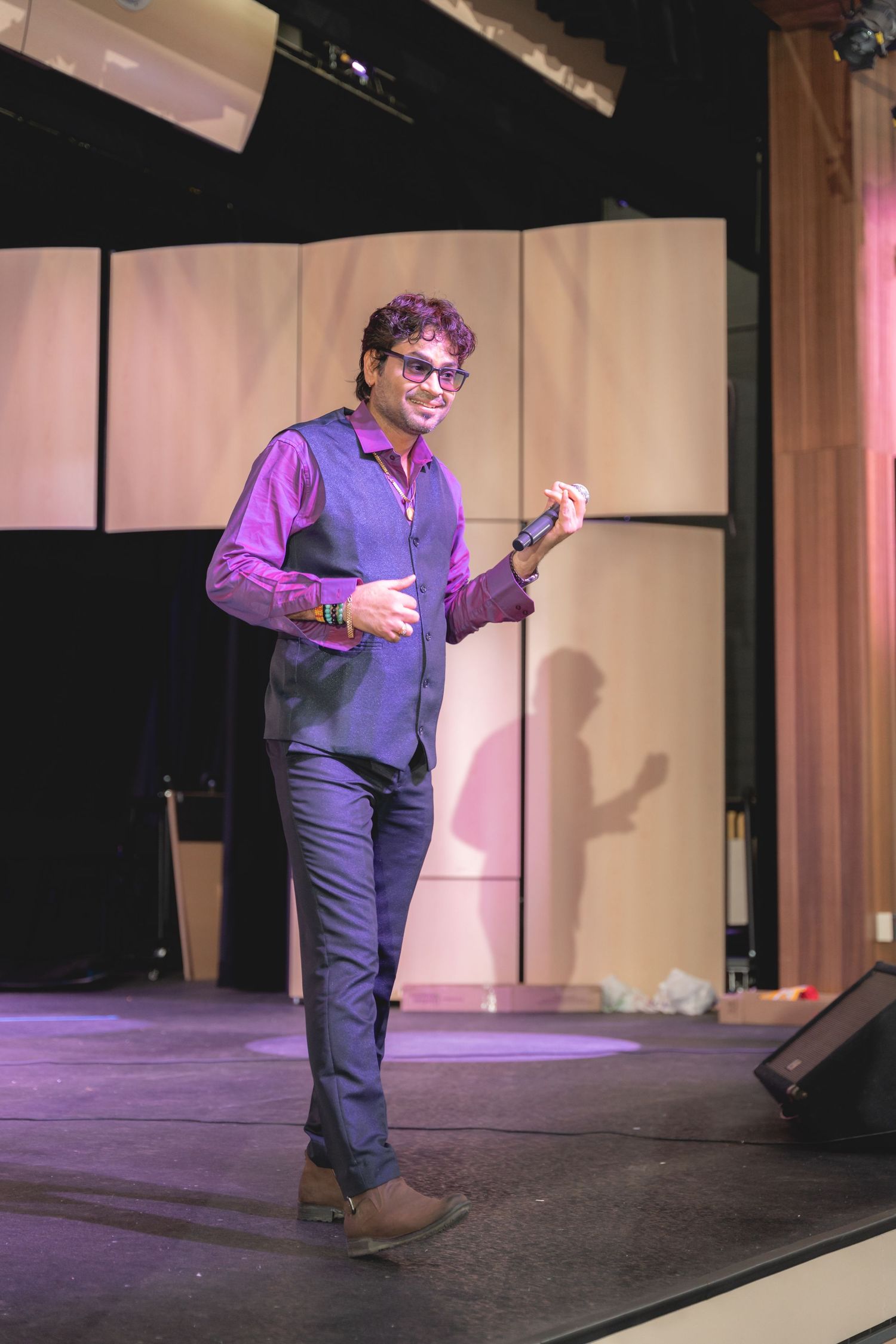 A performer in a purple shirt and dark pants gestures while standing on stage under theatrical lighting.