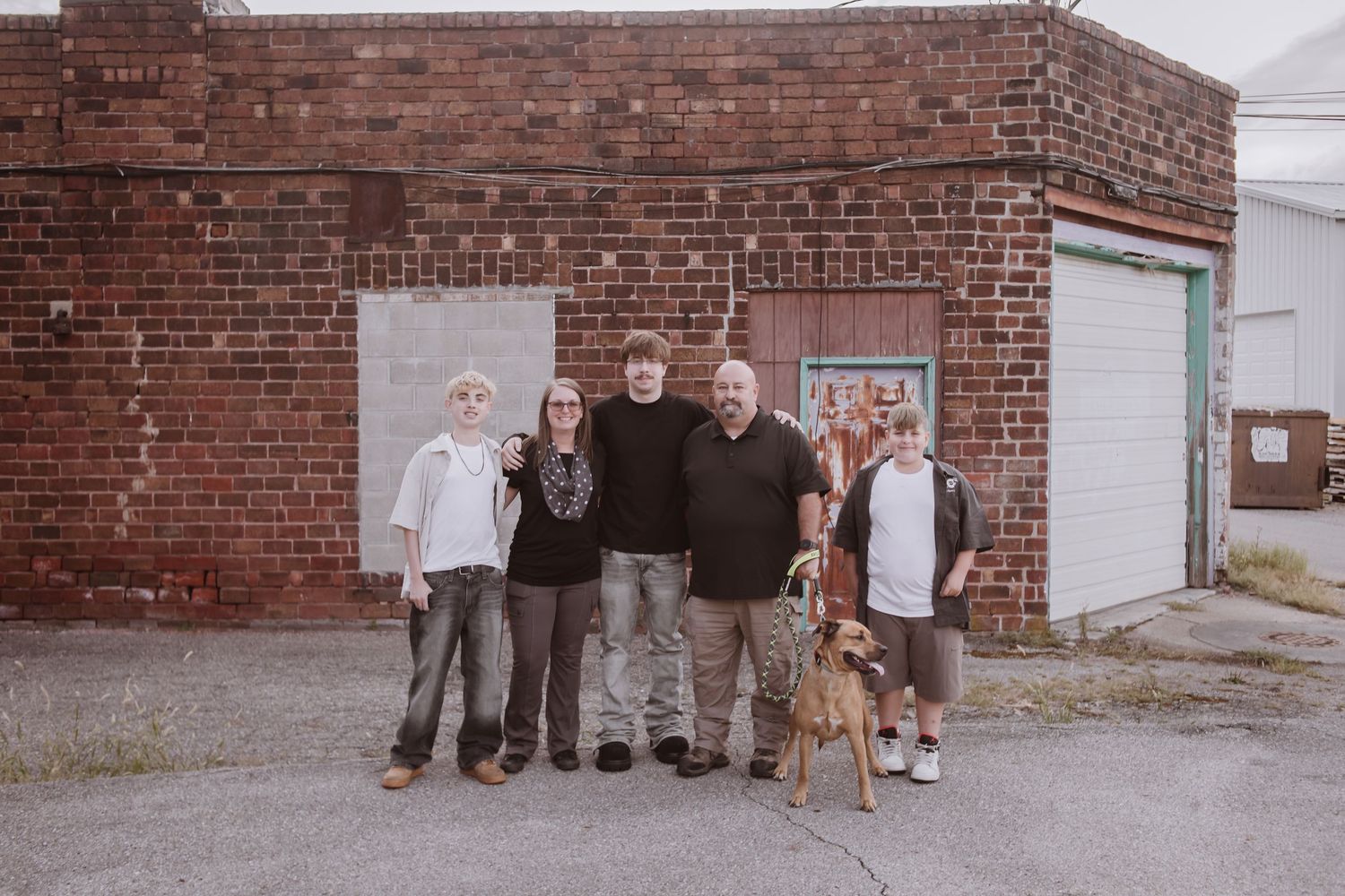 Family of four poses with their dog in front of a rustic red brick building outdoors.