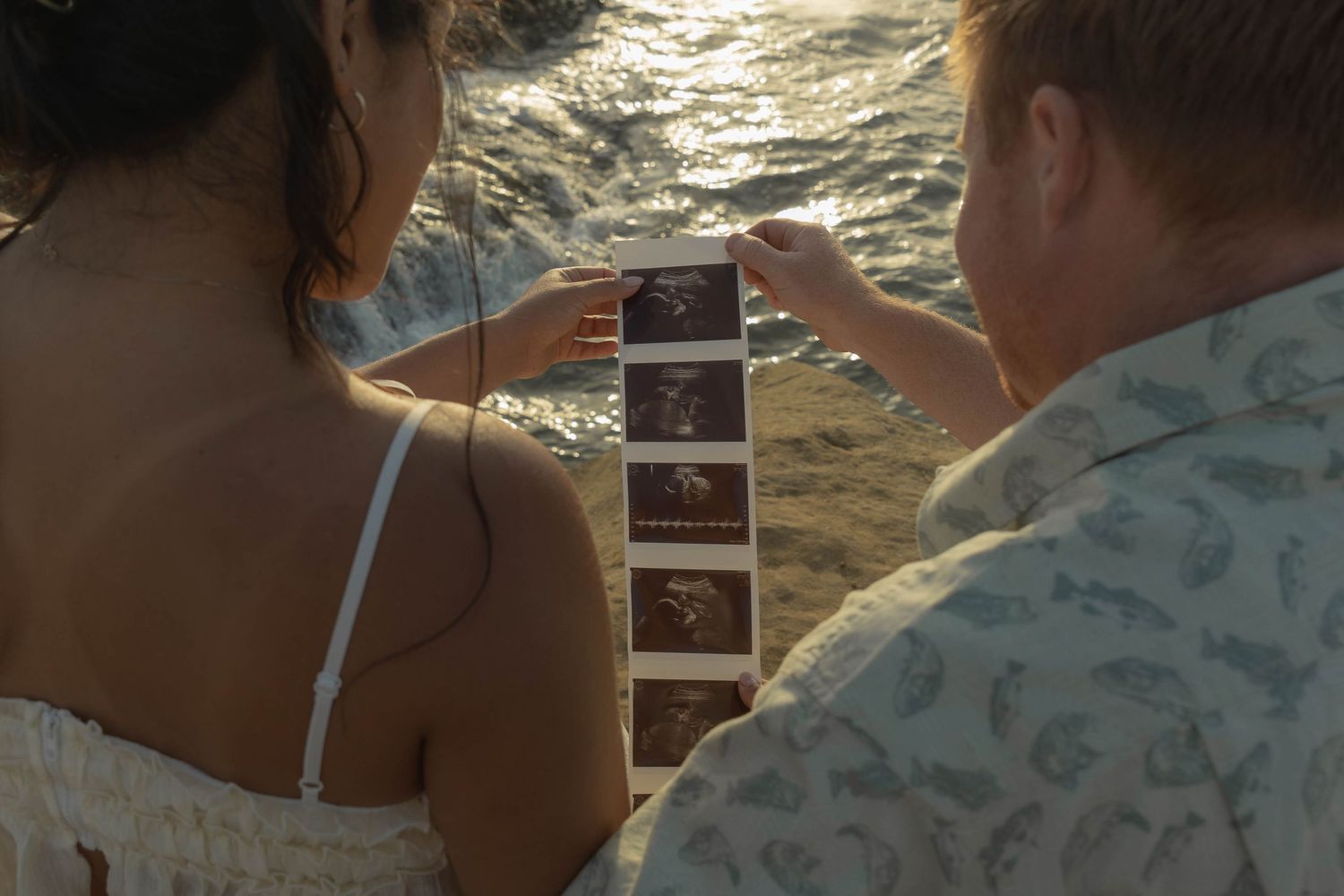 Two people look at photos together while sitting on rocks by the ocean.