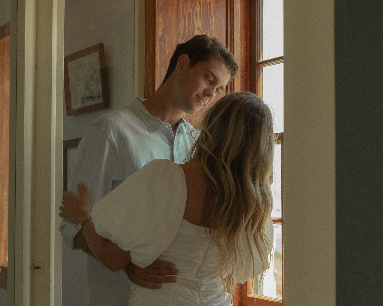 Romantic couple embracing and kissing near a sunlit window in a cozy room, woman in white dress.