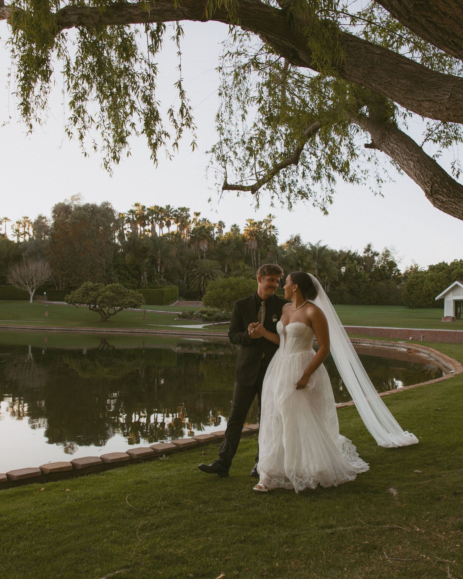 Bride and groom share a romantic moment under a willow tree beside a reflective pond at dusk.