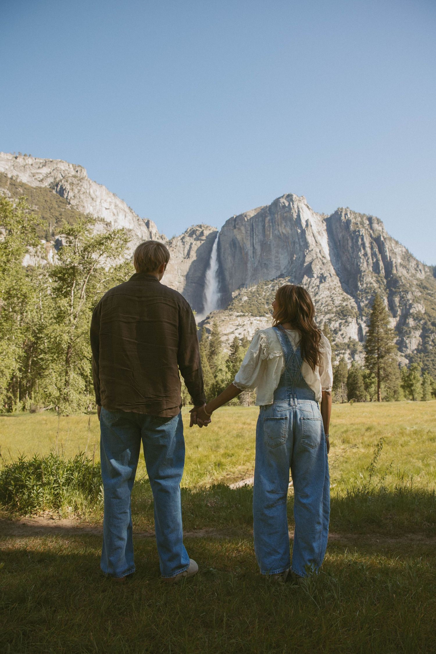 Couple holding hands facing Yosemite Falls waterfall with granite cliffs and green meadow on sunny day.