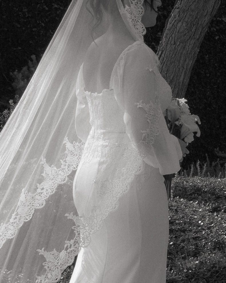 Black and white photo of a bride in a lace wedding gown and veil, holding a bouquet outdoors.