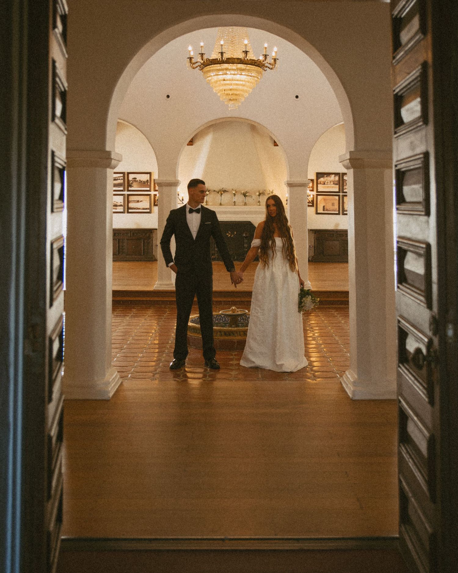 Elegant wedding couple holding hands under a chandelier-lit archway in a grand historic venue.