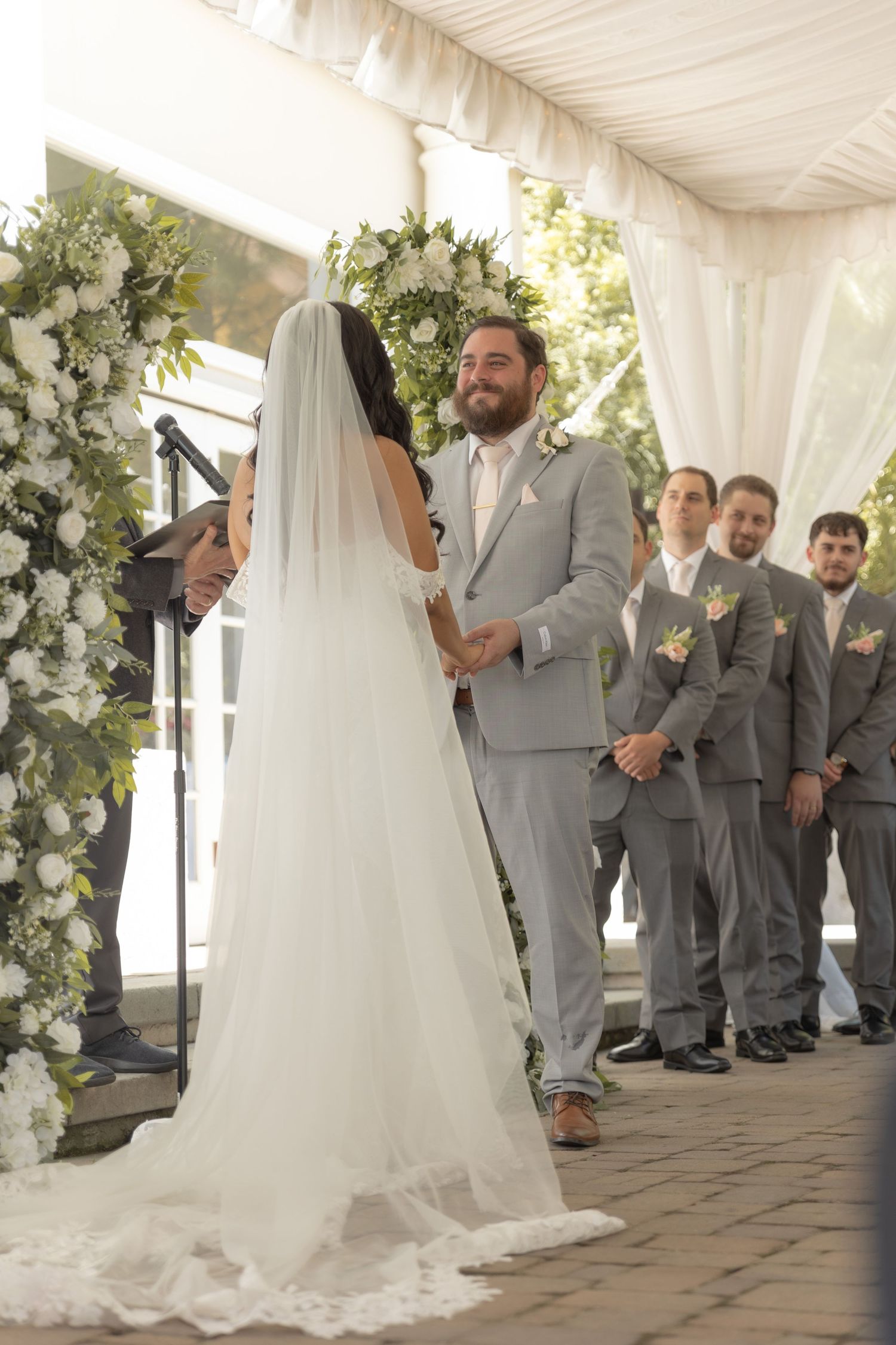 Wedding ceremony under white tent with floral arch and wedding party in grey suits.