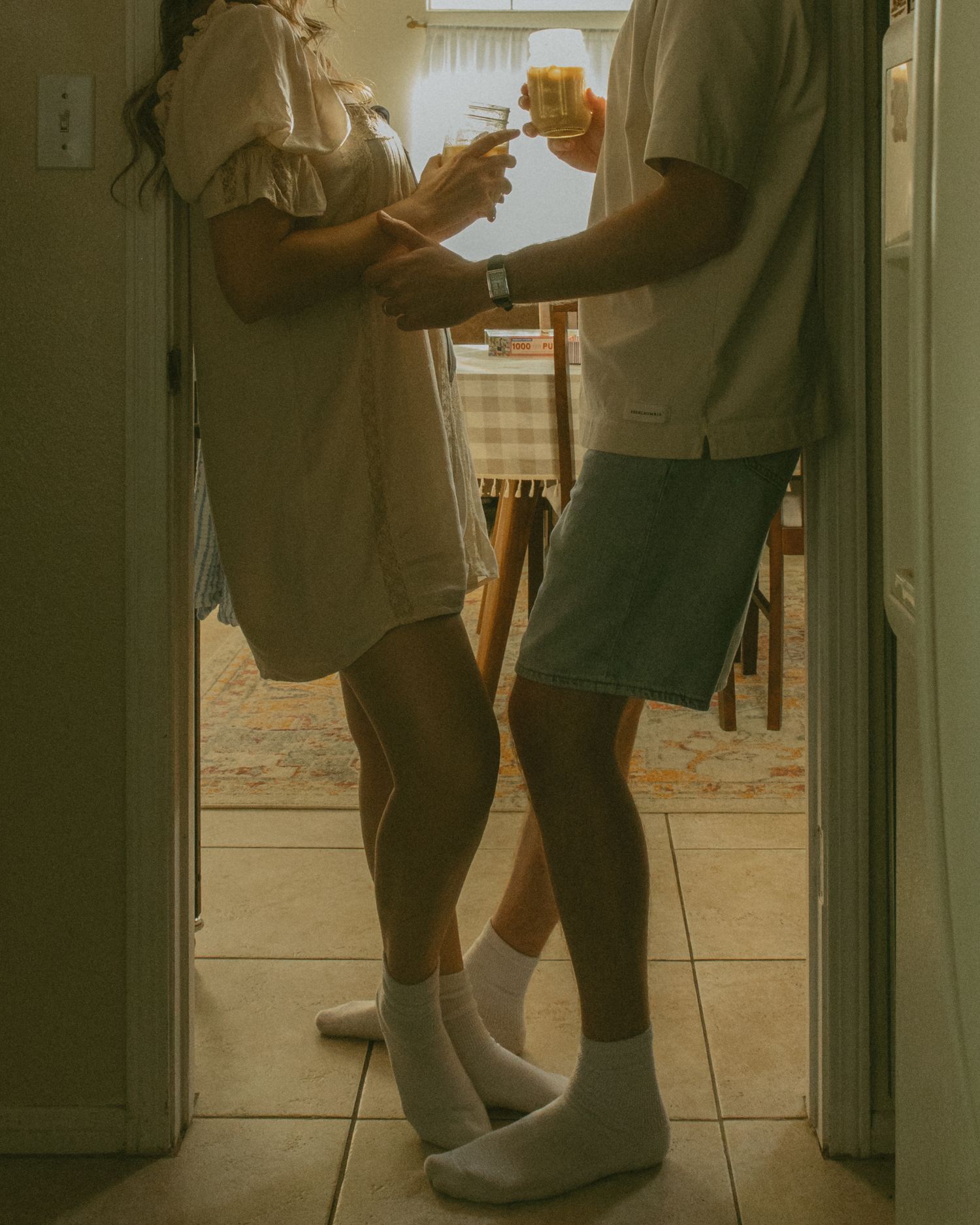 Couple standing in doorway clinking glasses with orange juice, wearing casual summer outfits and white socks on tile floor.