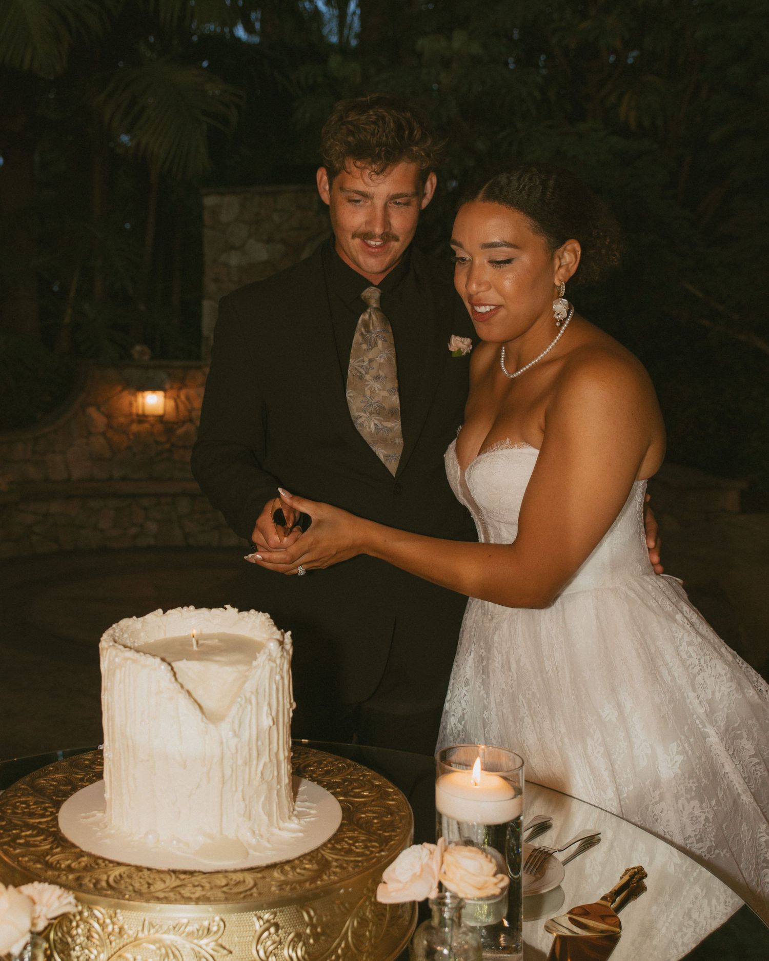 Bride and groom cut white wedding cake together at elegant outdoor evening wedding reception.