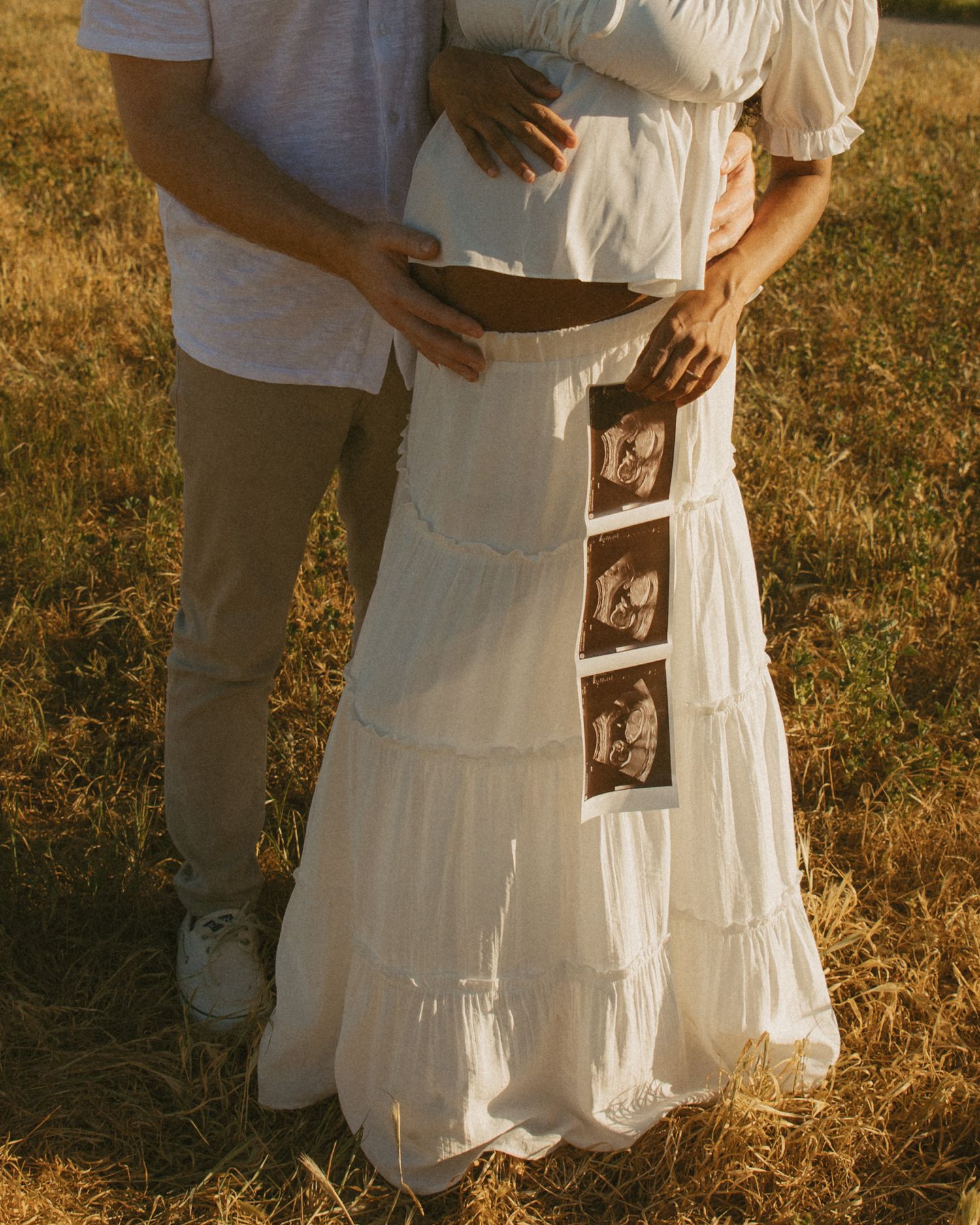 Couple holding ultrasound photos in a sunny field, woman in white dress with visible baby bump, pregnancy announcement.
