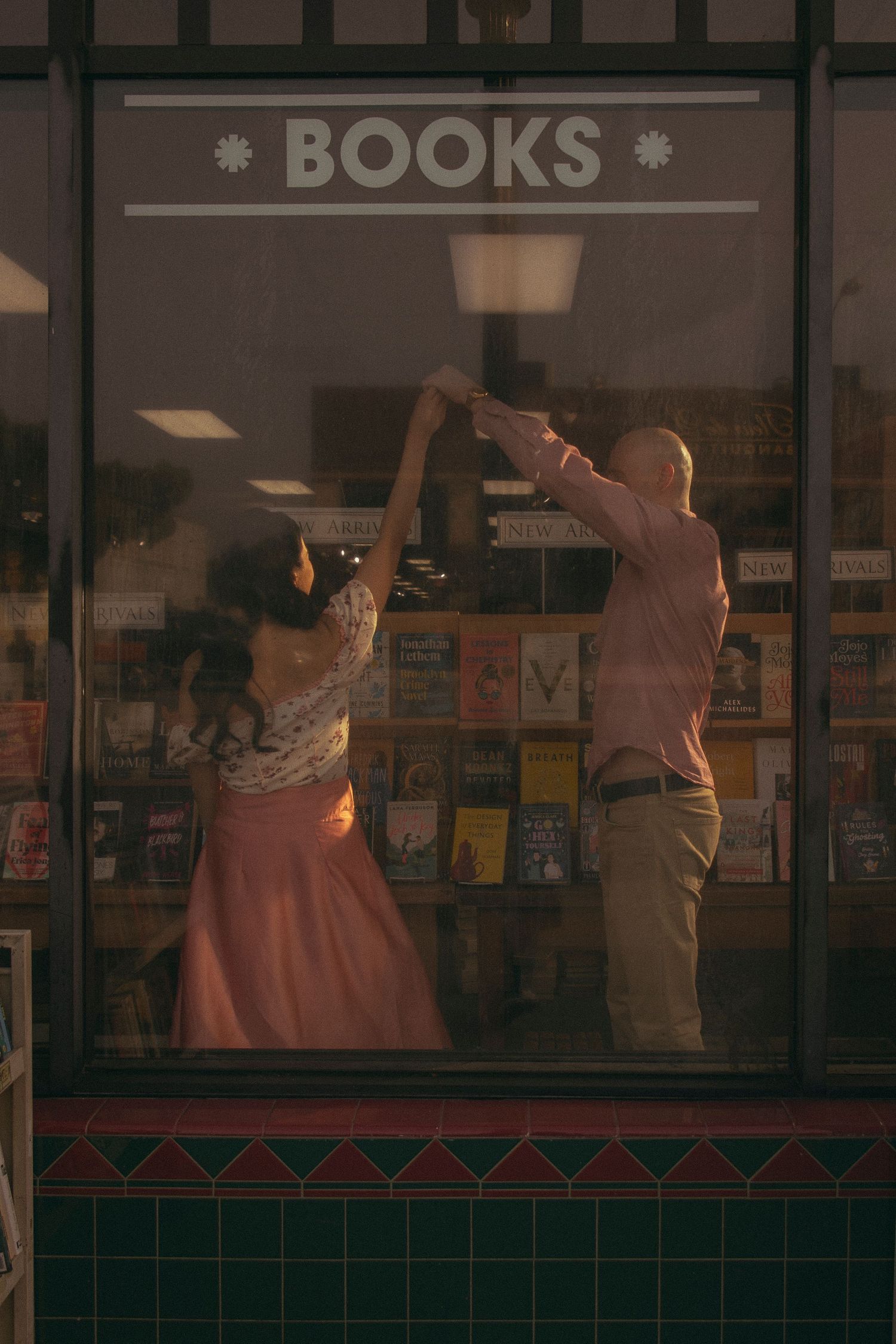 Couple dancing in front of a bookstore window display at golden hour, viewed from outside.