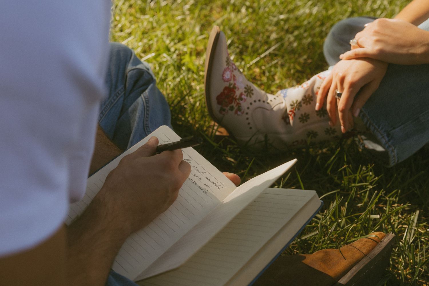 Two people sitting on grass outdoors, one writing in a notebook, the other wearing decorative cowboy boots.