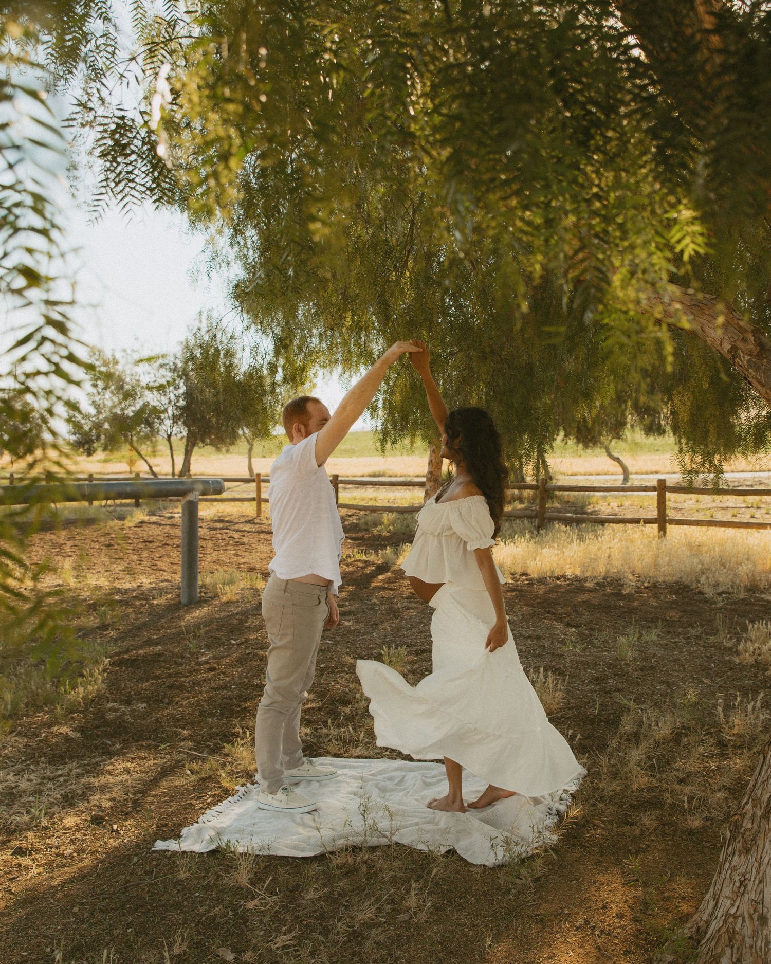 Couple dancing outdoors under a willow tree in white outfits on a sunny day in a rustic farm setting.