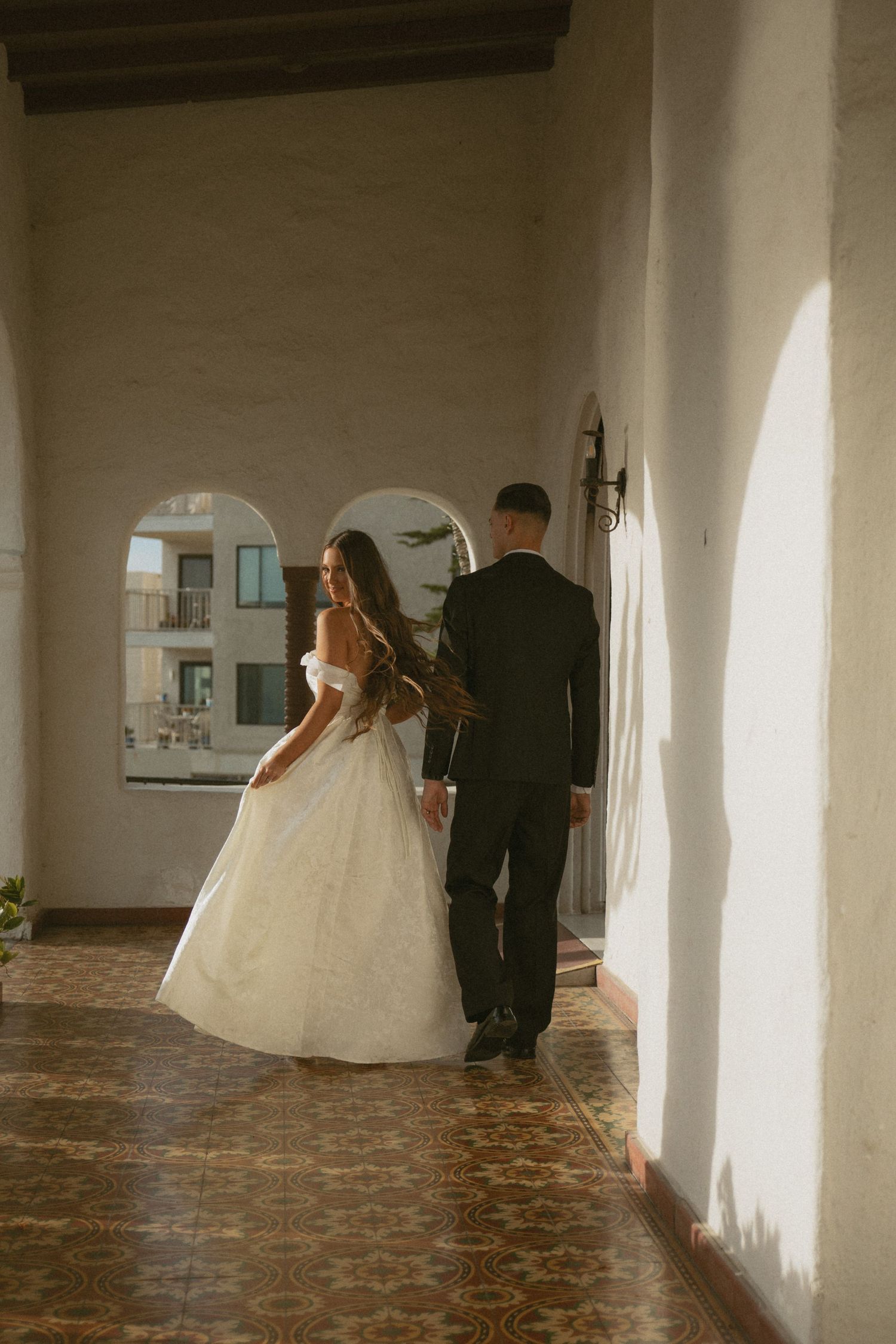 Bride and groom walk through sunlit arched corridor with ornate tile floors at wedding venue.