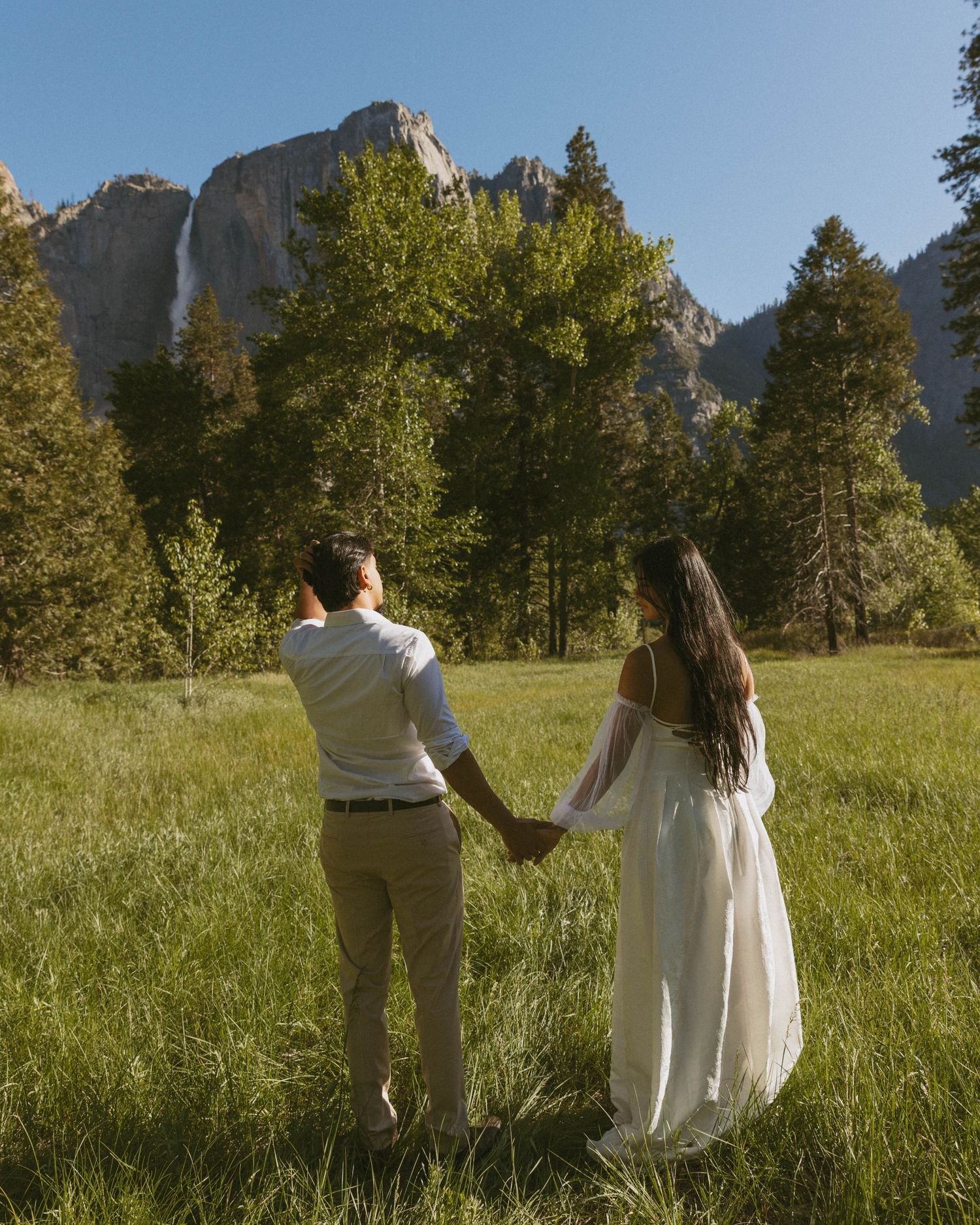 Couple in wedding attire holding hands in Yosemite meadow with waterfall and granite cliffs in background.