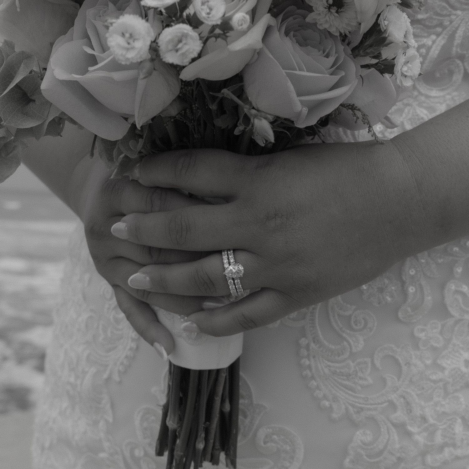Close-up black and white photo showing hands holding a bridal bouquet with roses and lace details.