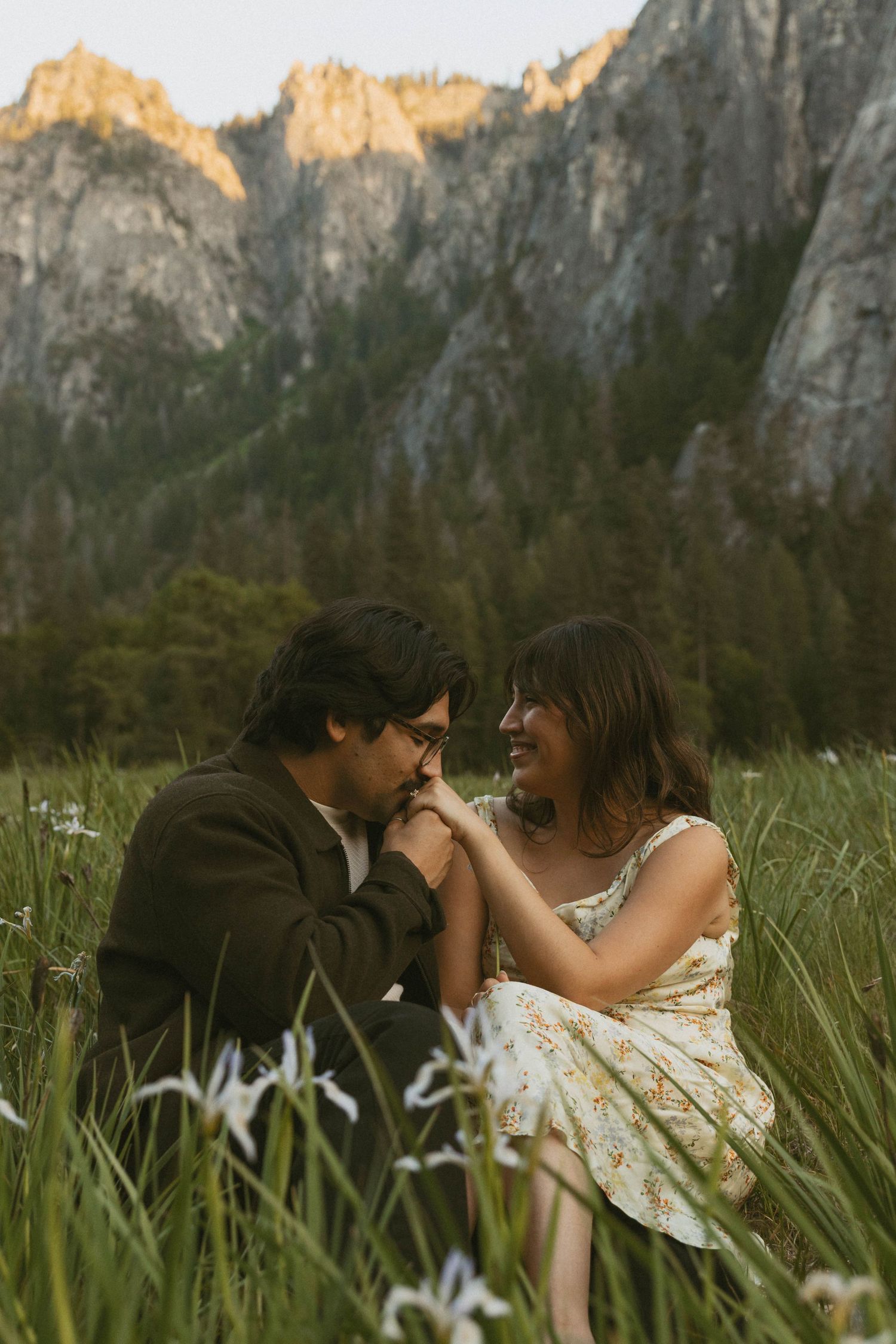 Couple sitting in a wildflower meadow holding hands, smiling at each other with rocky mountains and trees in background.