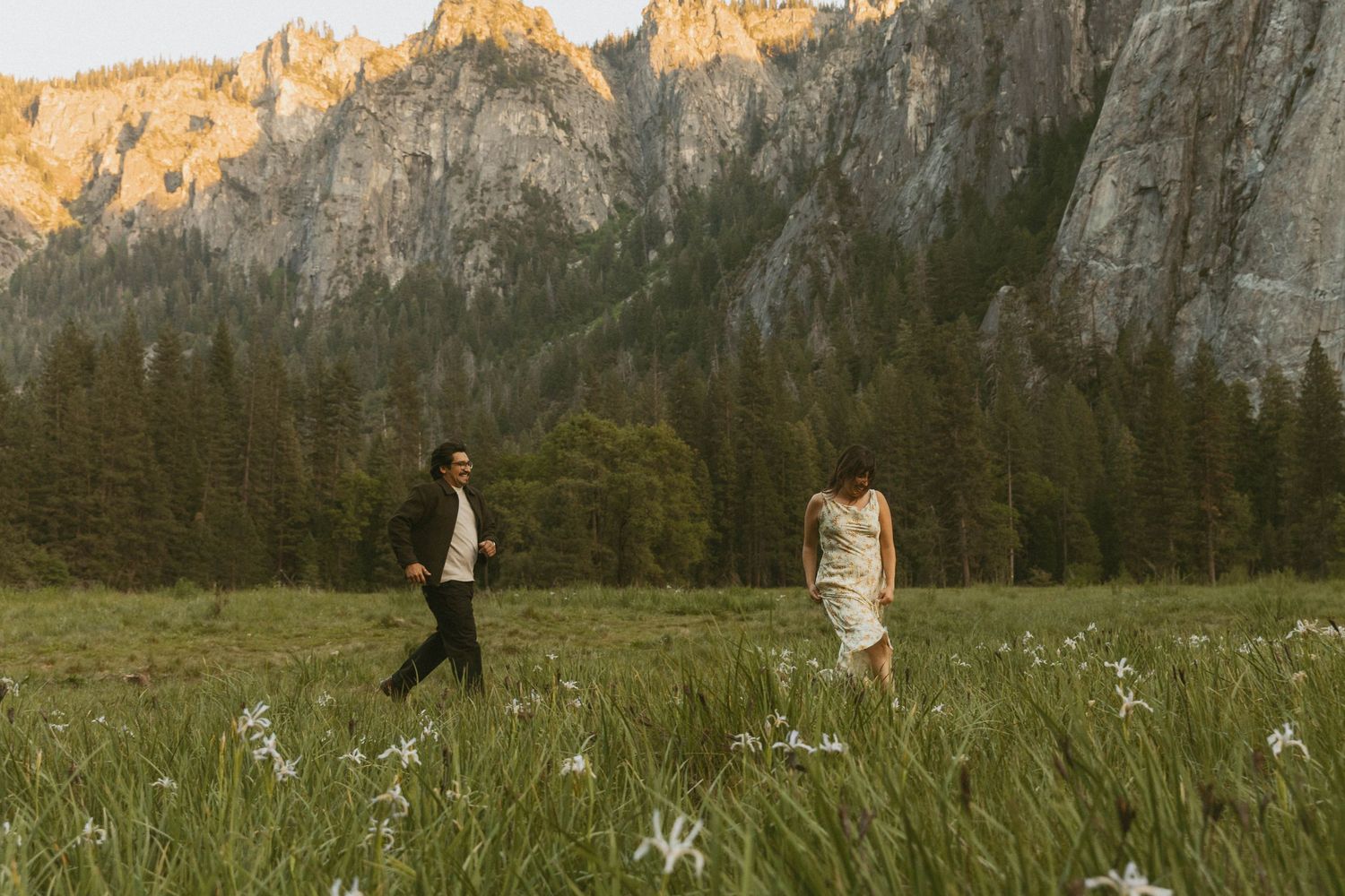 Couple walking through wildflower meadow with dramatic granite cliffs and pine forest backdrop at golden hour.