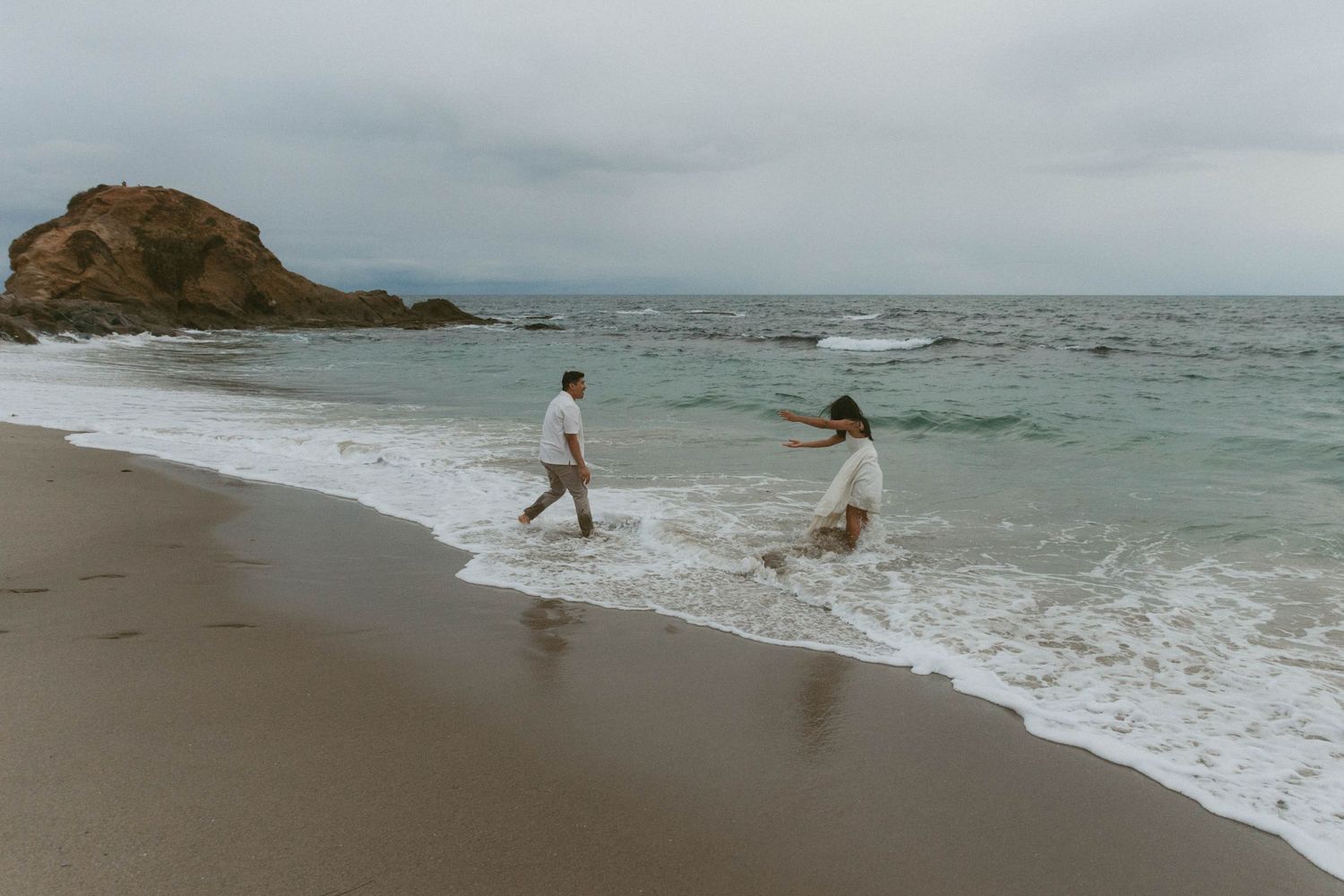 Couple playing in ocean waves on a rocky beach during an engagement photo session on an overcast day.