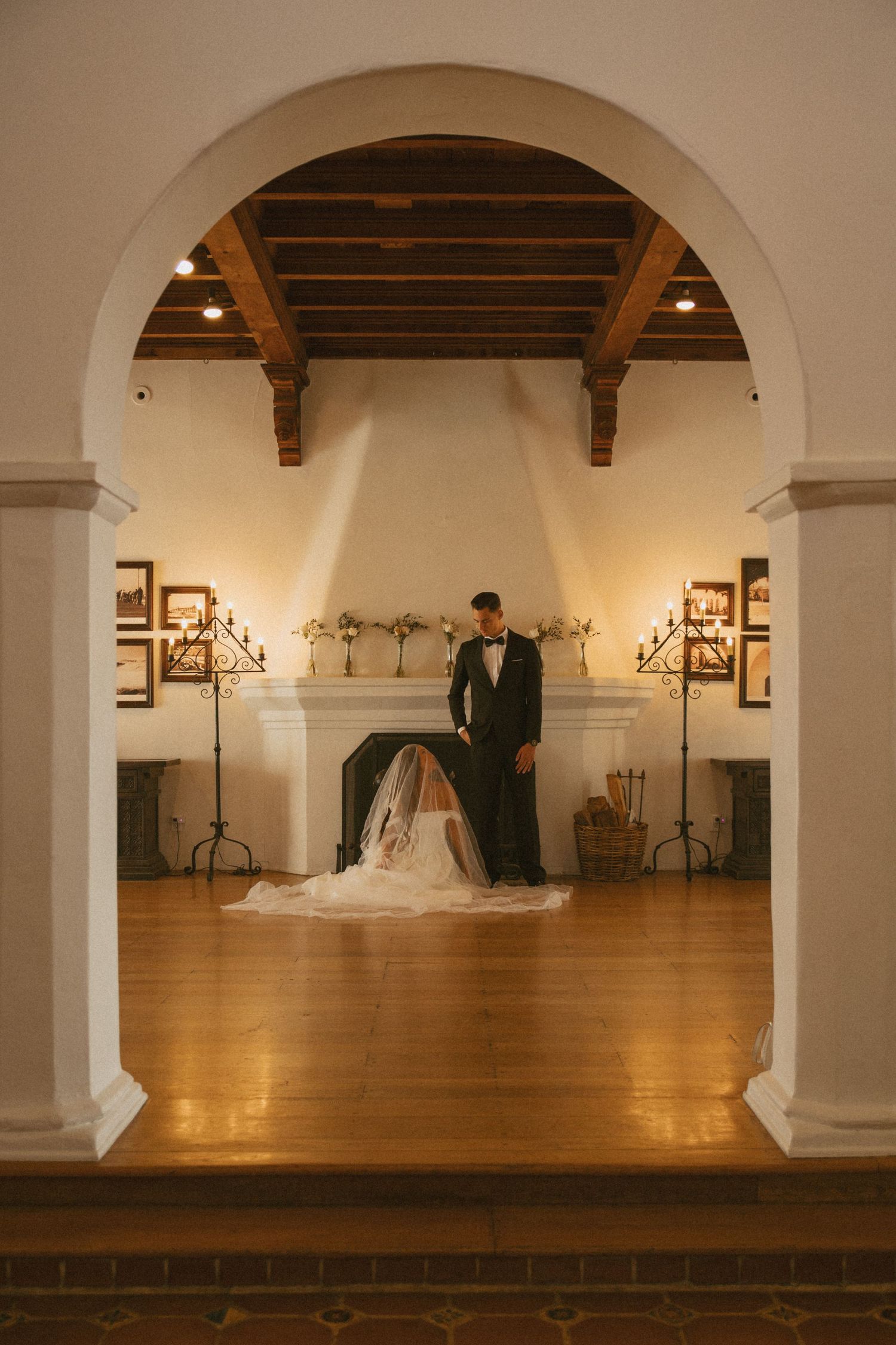 Bride and groom framed by elegant arch near fireplace in warmly lit wedding venue with wood beam ceiling.