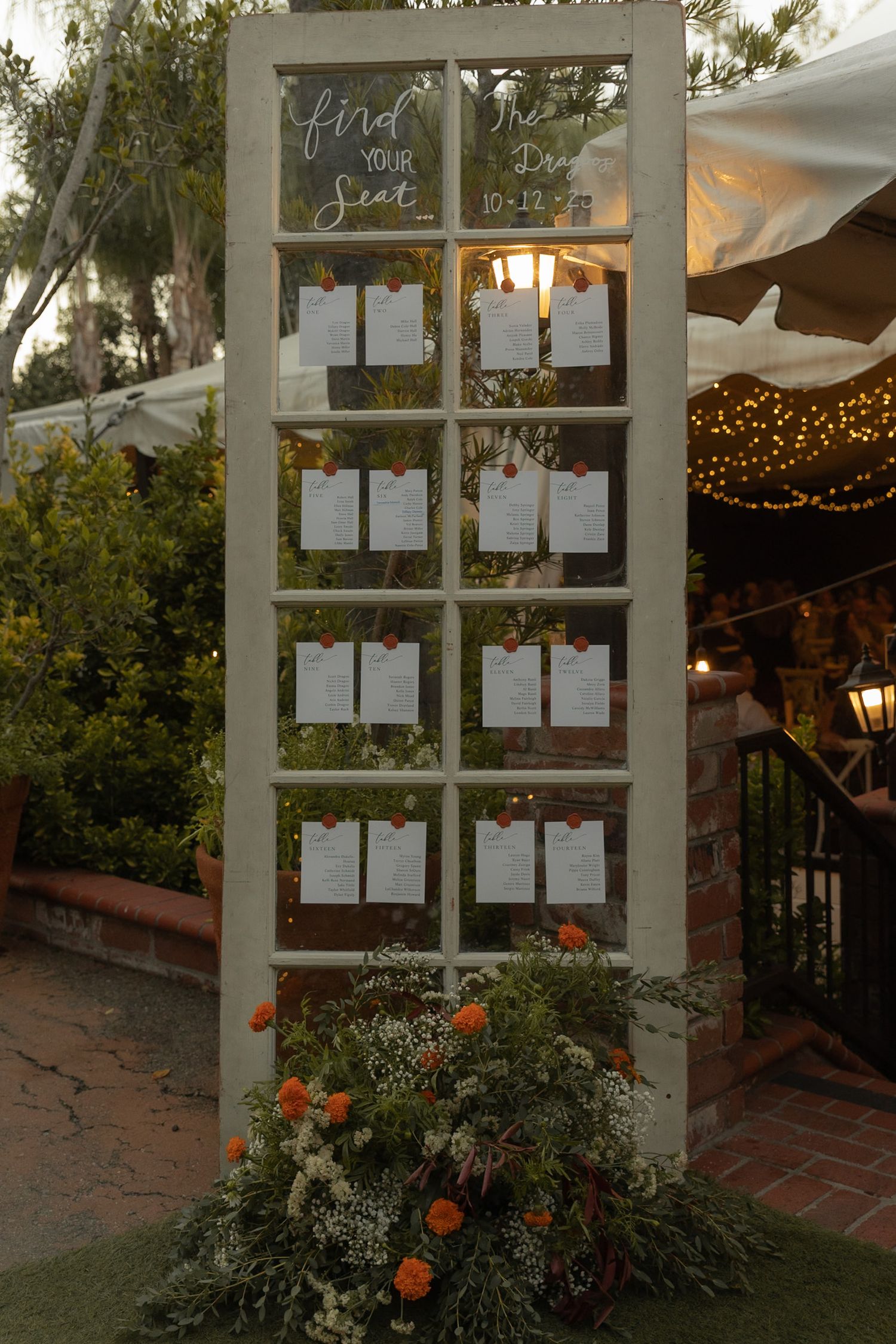 Rustic wedding seating chart displayed on a vintage white door frame with orange floral arrangements.