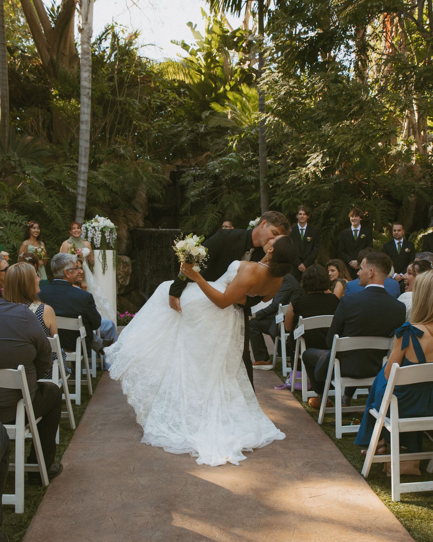 Bride and groom kiss on aisle after outdoor garden wedding ceremony surrounded by seated guests.