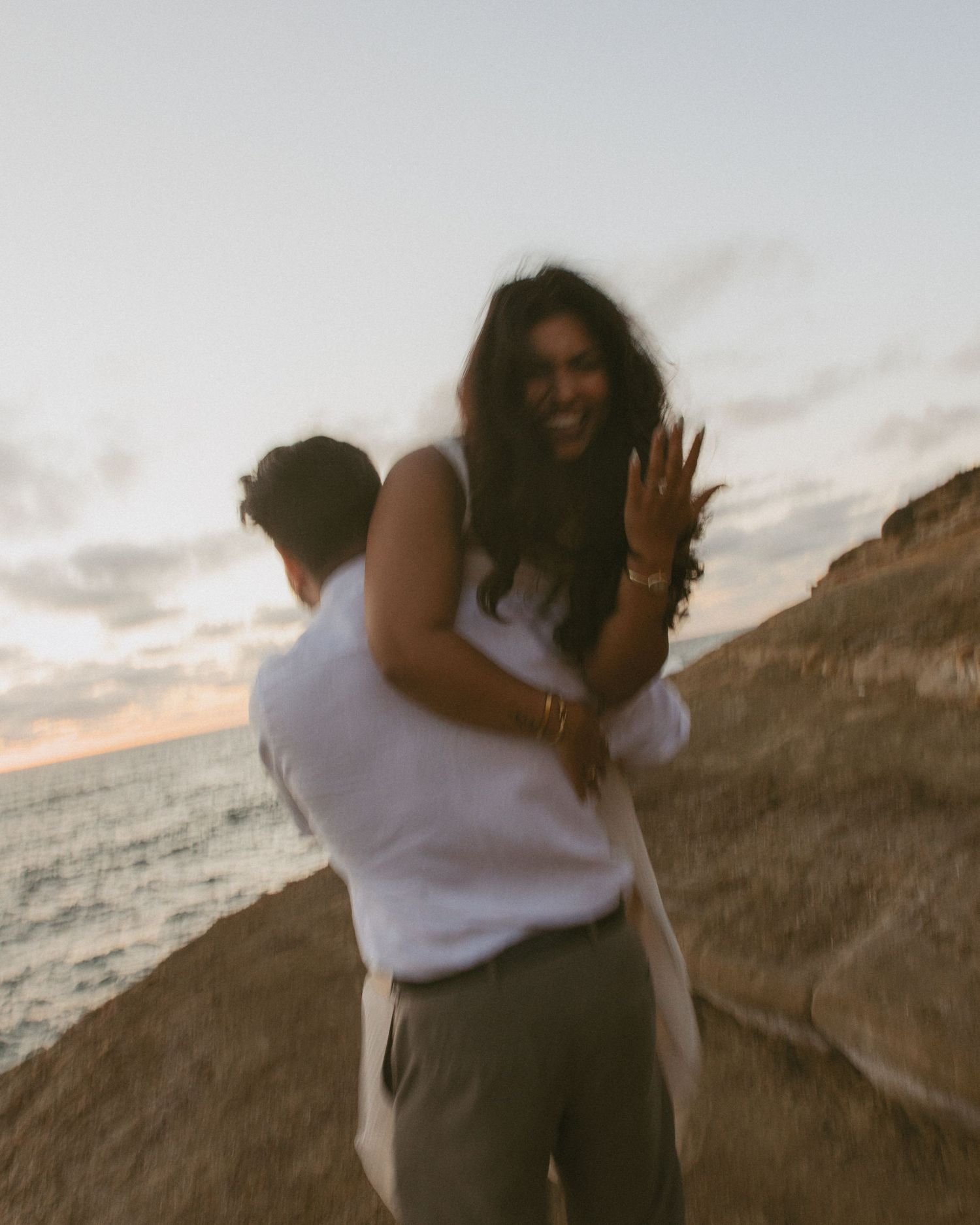 Couple embracing on a cliffside at sunset, woman smiling and showing engagement ring with ocean in background.
