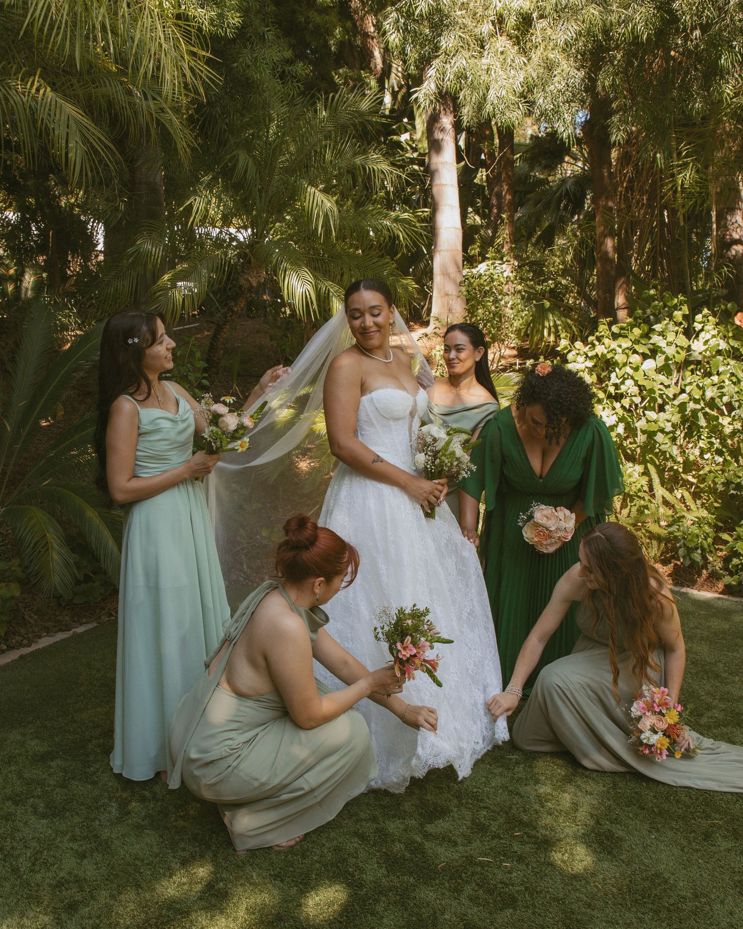 Bride in white gown surrounded by bridesmaids in sage green dresses in lush garden setting.
