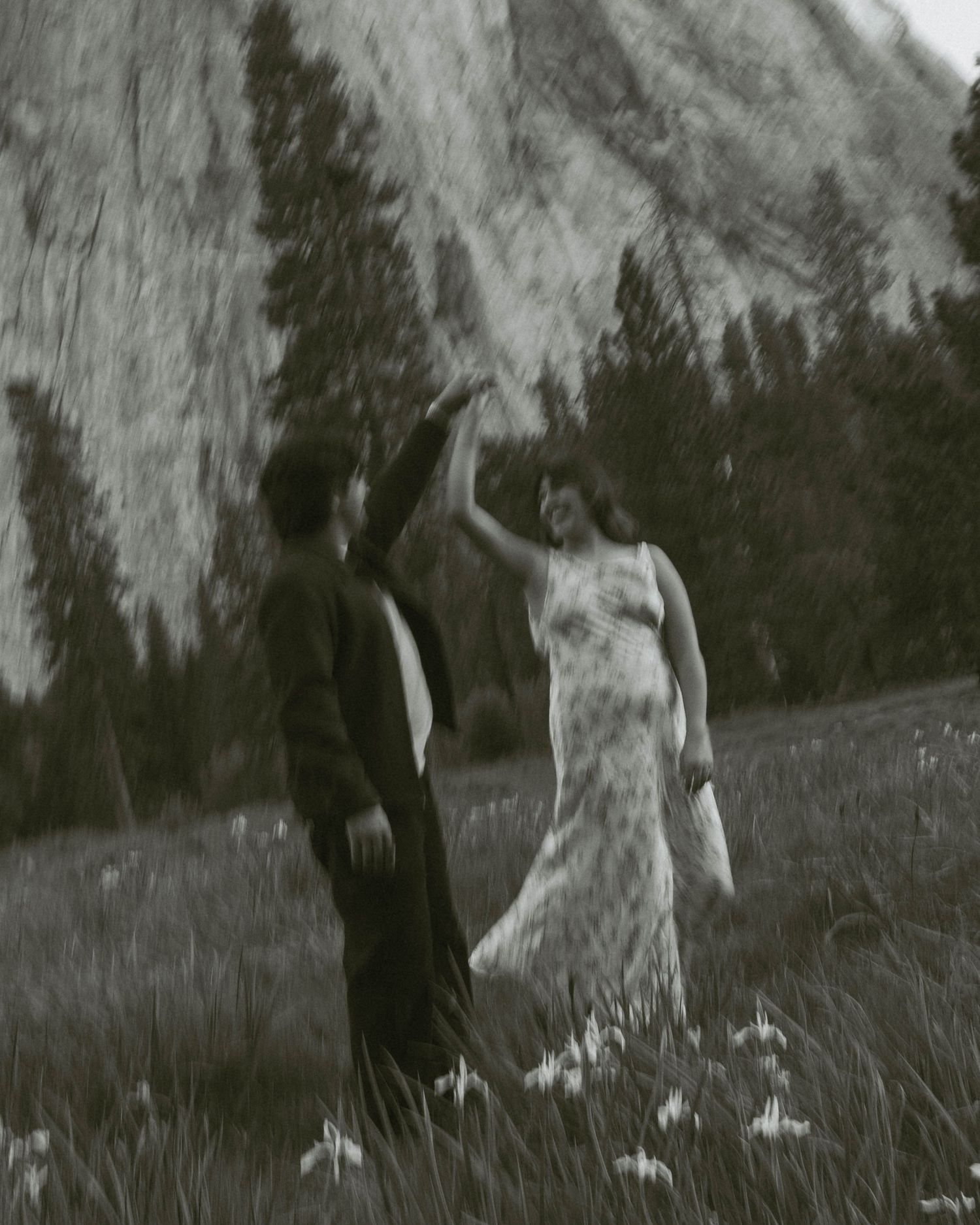 A couple dances in a meadow with wildflowers, framed by towering granite cliffs and pine trees in a vintage black-and-white photo.