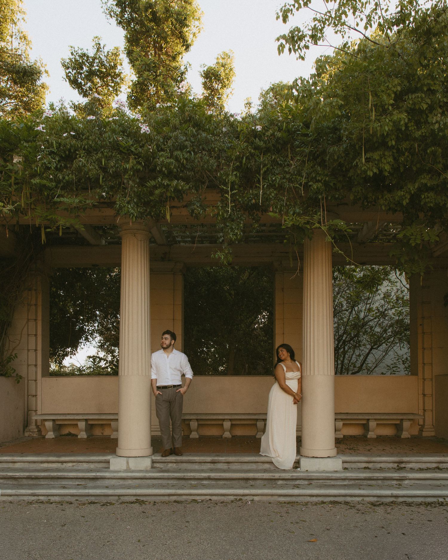 Couple poses between classical columns draped in greenery at an elegant outdoor pergola during golden hour.