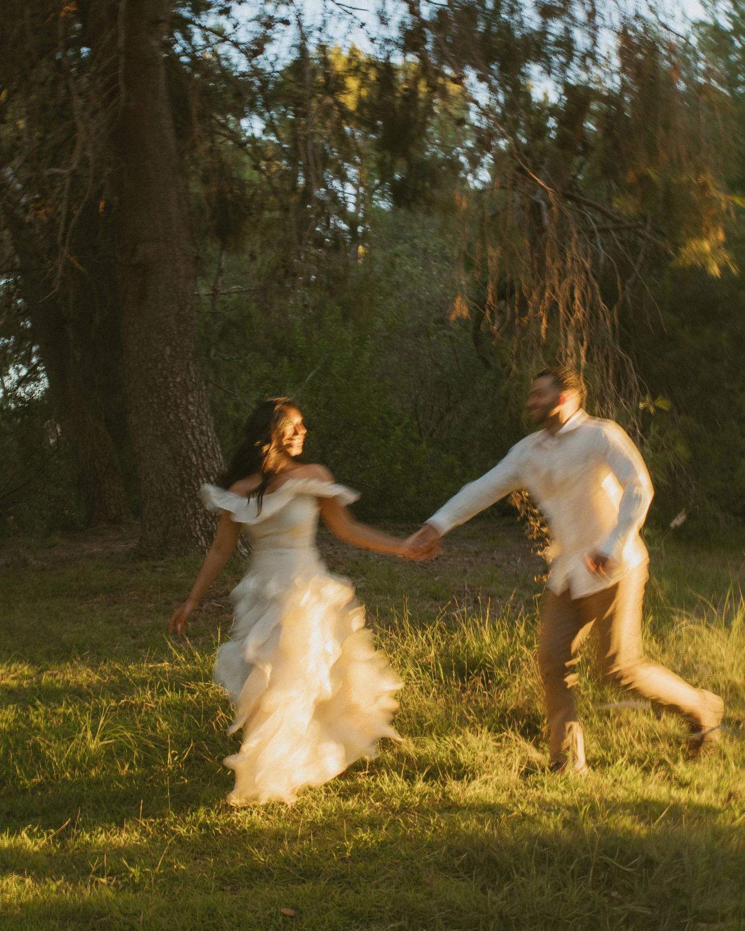 A bride in a flowing white gown and groom run hand-in-hand through a sunlit forest clearing.
