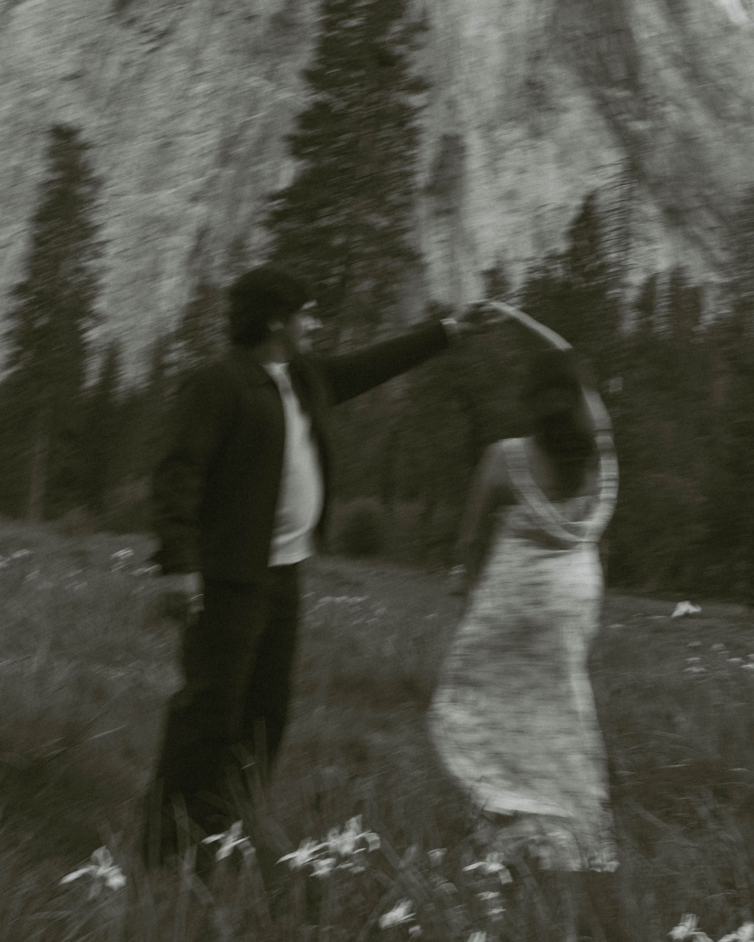 Black and white photo of a couple dancing in a mountain meadow surrounded by pine trees and a rocky cliff.