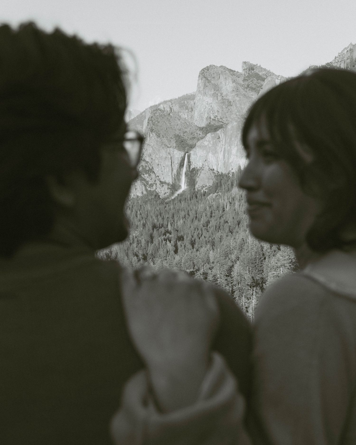 Black and white photo of a couple facing each other with Yosemite's Half Dome and forest visible in the background.