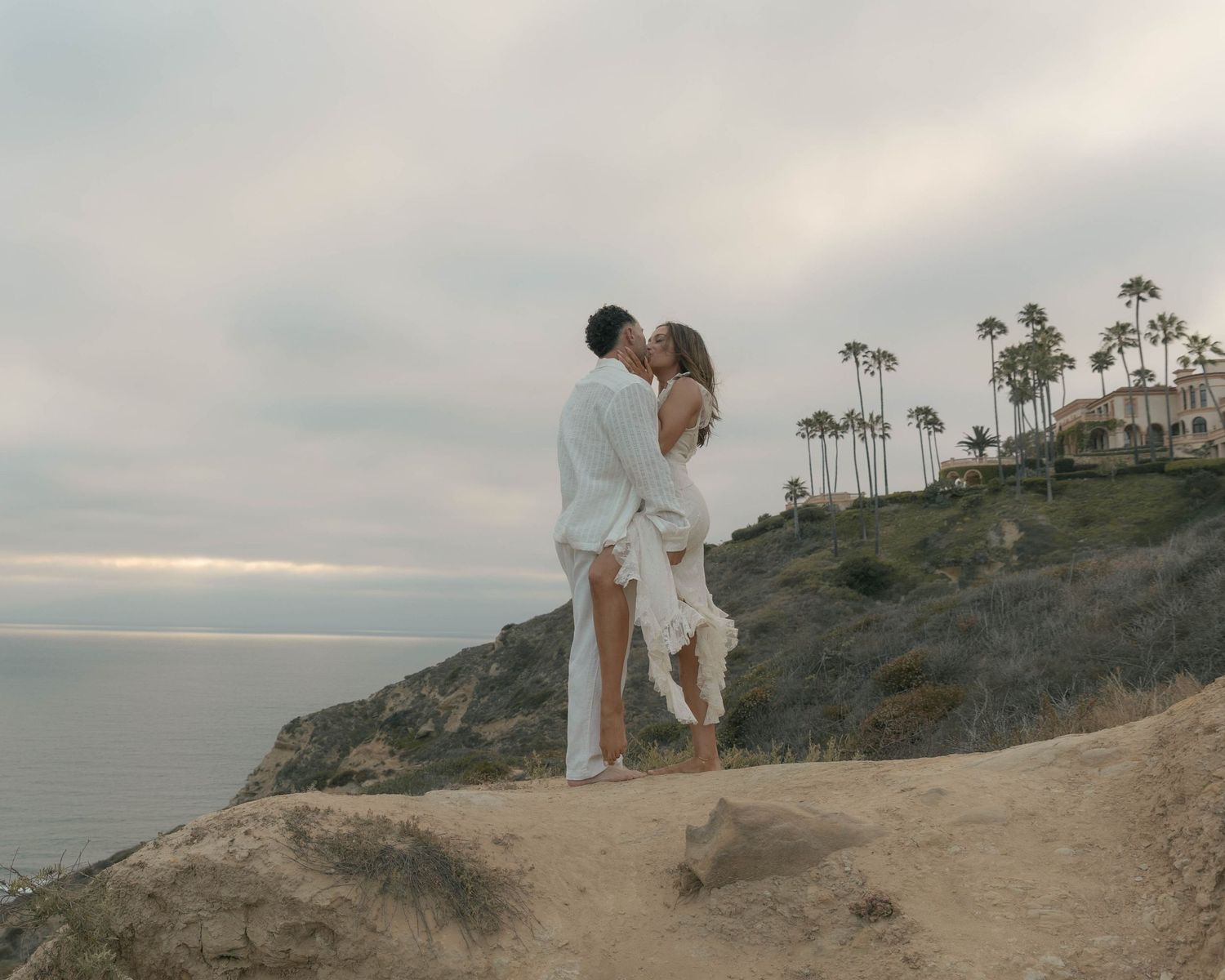 A couple in white clothing walks along a scenic coastal cliff path.