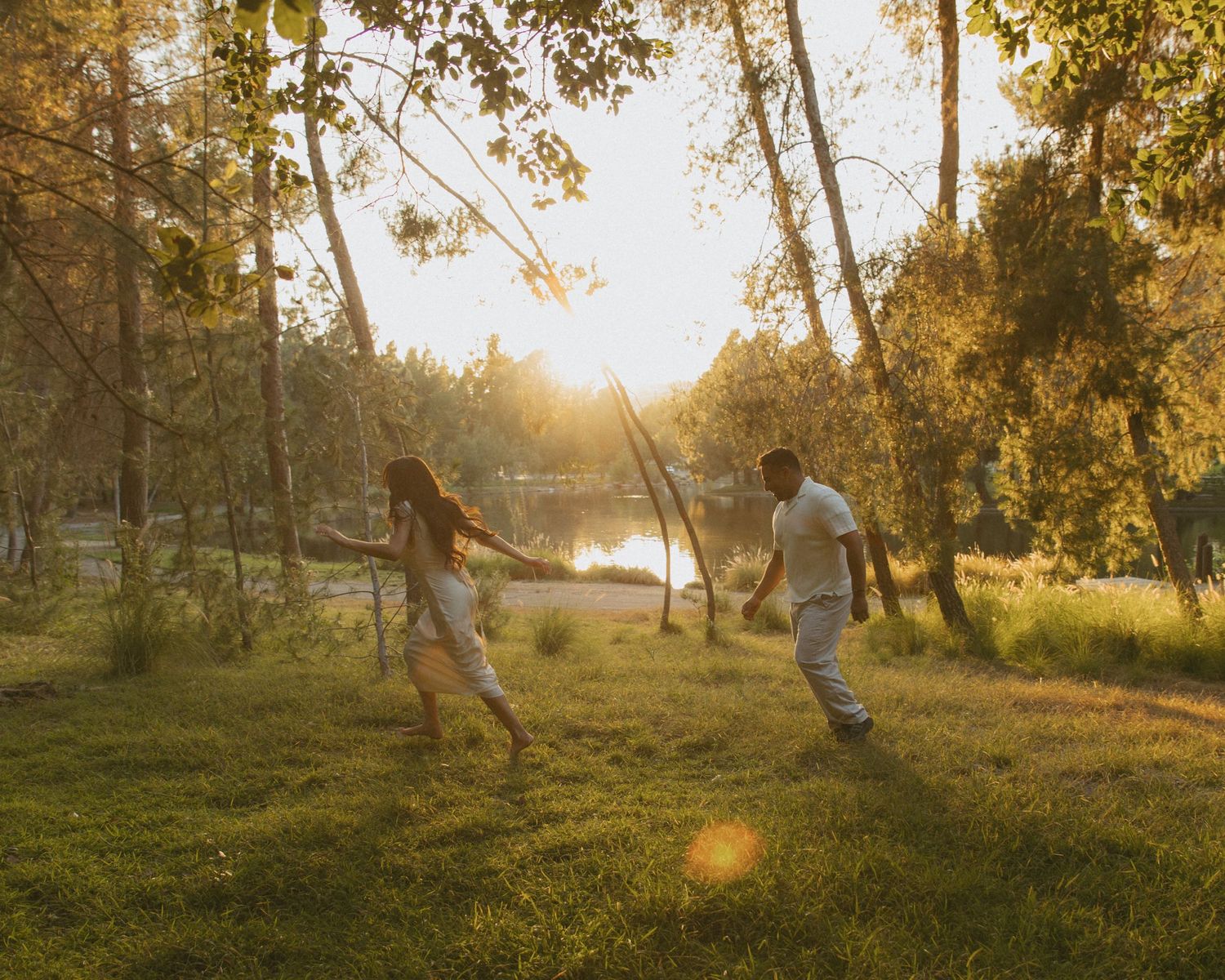Couple dancing playfully in a sunlit forest clearing near a lake during golden hour sunset.