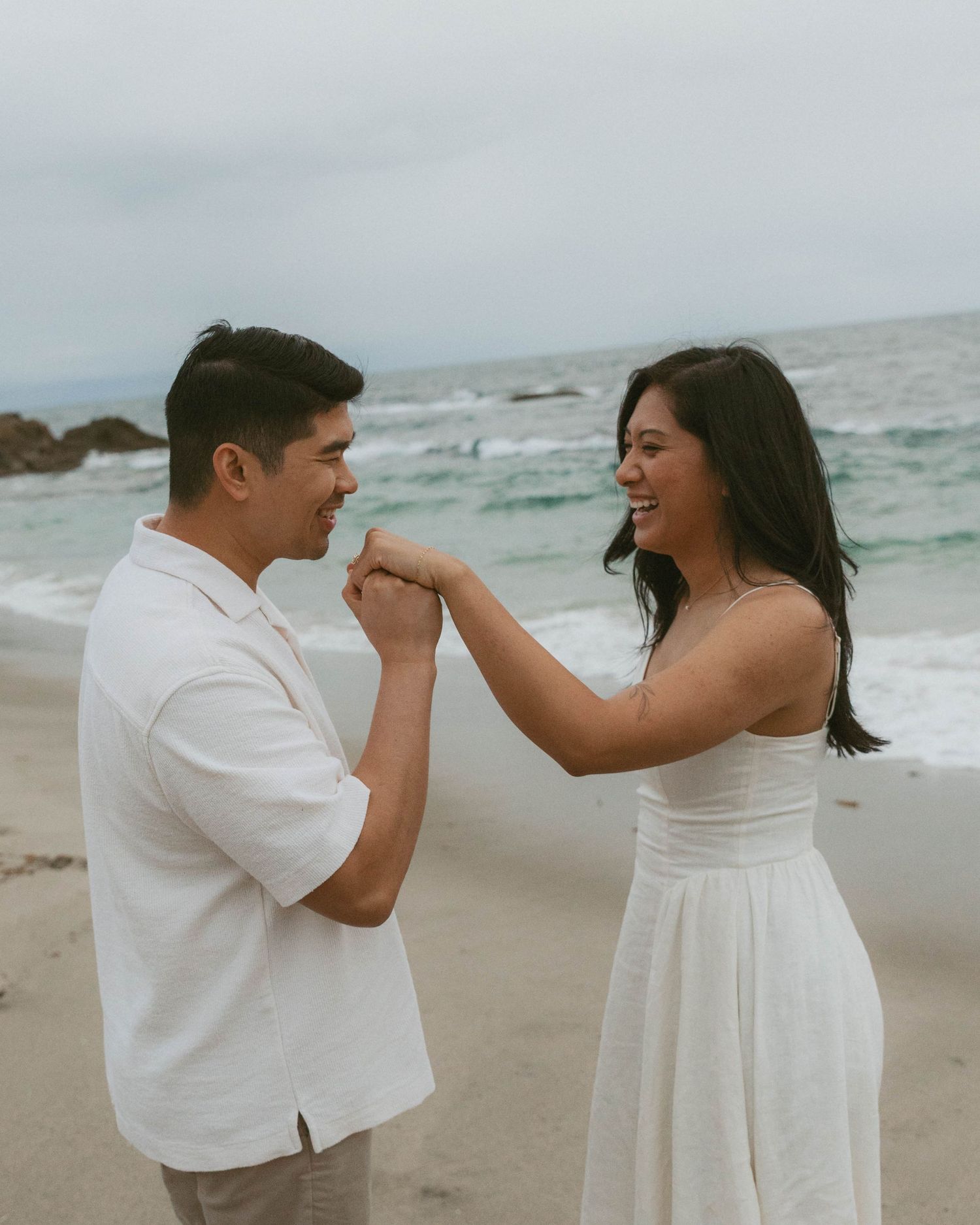 A couple in white outfits shares a romantic moment on a beach, the man kissing the woman's hand as she smiles.