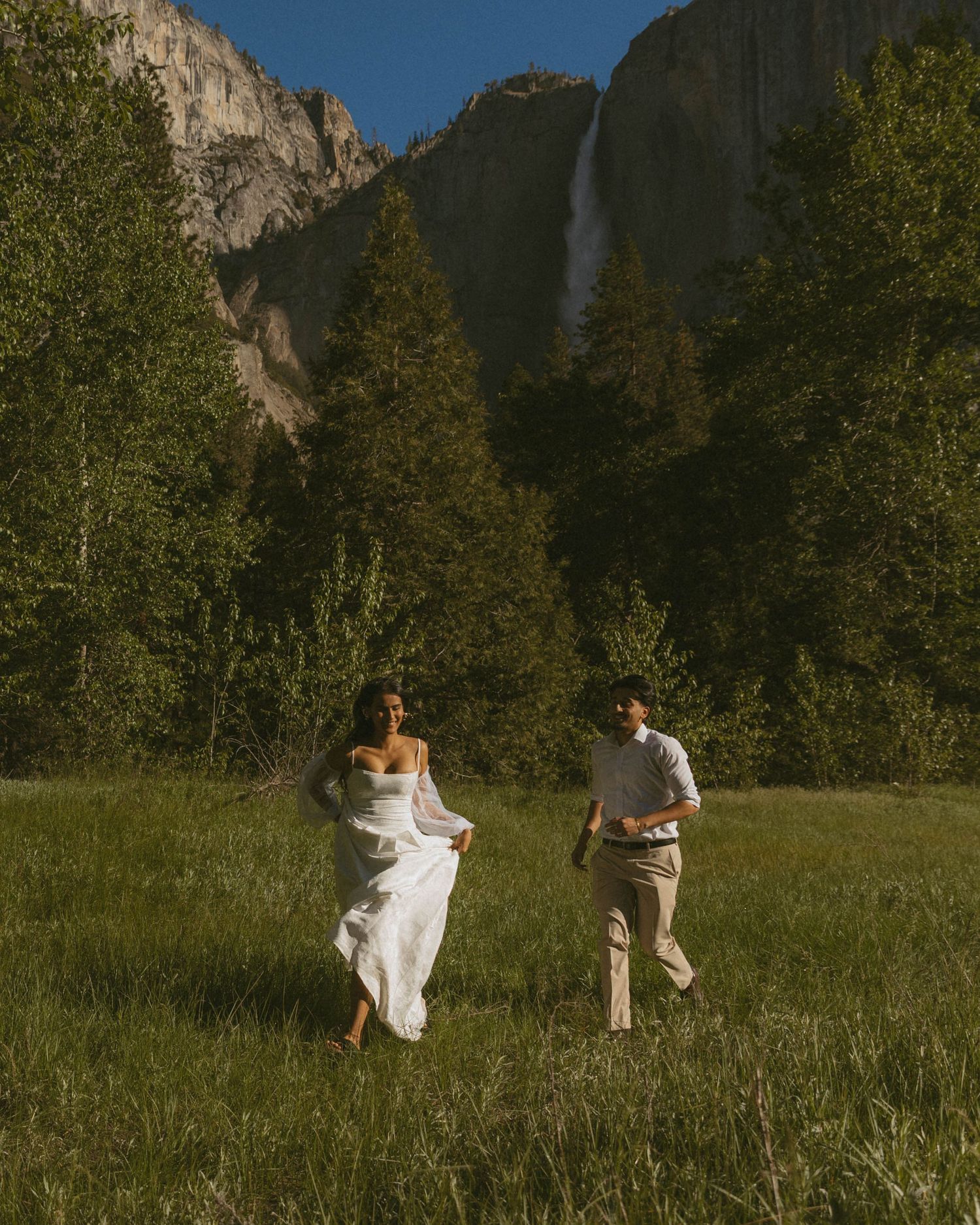 Couple in wedding attire running through a sunlit meadow with Yosemite Falls and tall trees in background.
