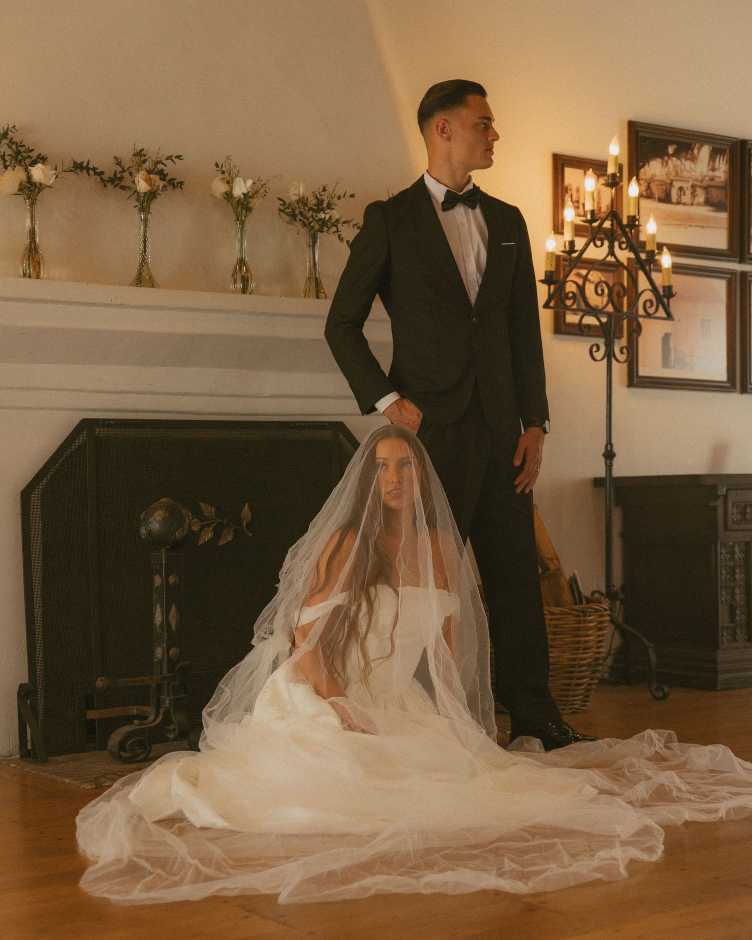 Elegant bride sitting on the floor in a long veil beside groom in black tuxedo near a fireplace.