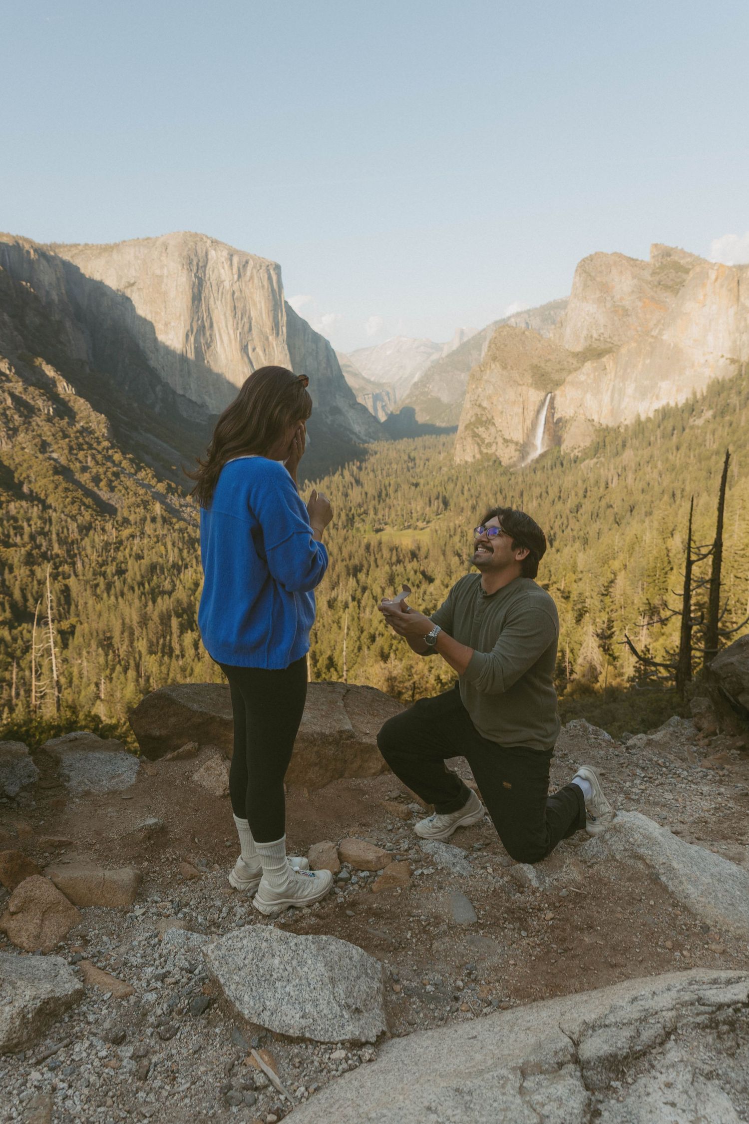 Man proposes to woman on one knee at Yosemite Valley overlook with El Capitan and Bridalveil Fall in background.