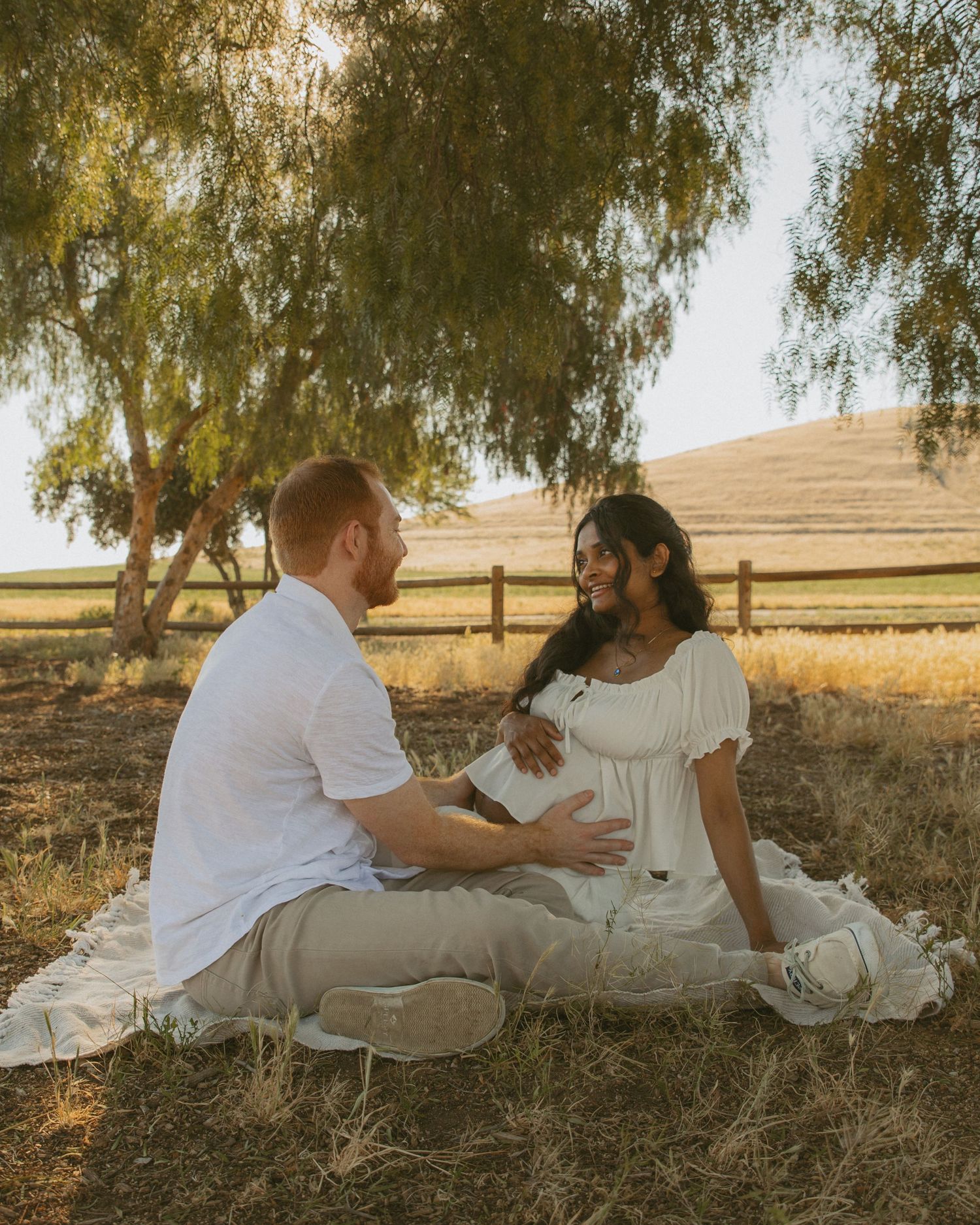 A couple sits on a blanket under trees during a golden hour maternity photo shoot in a rustic field.