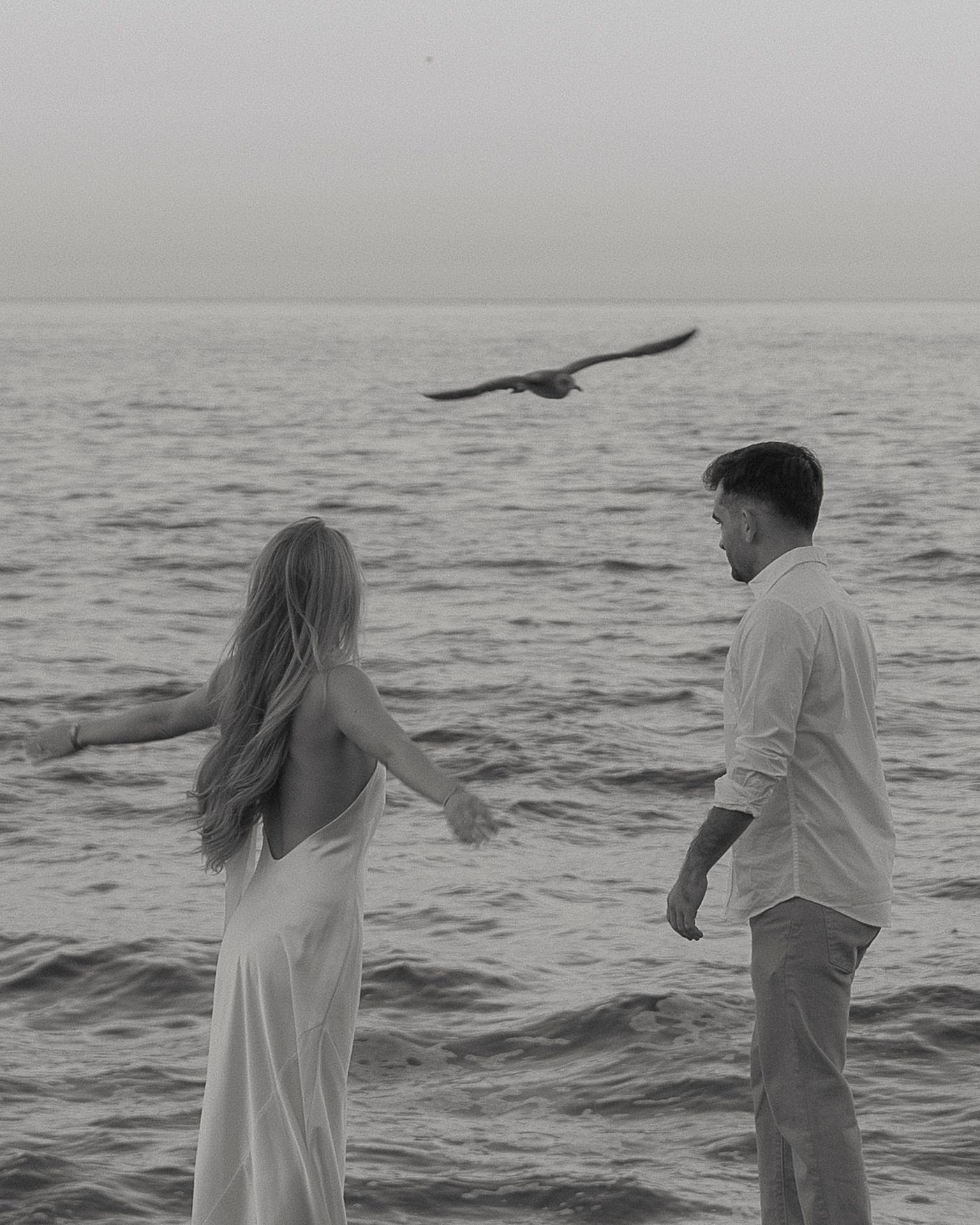 A couple in white and light colored clothing stands by the ocean while a bird flies overhead in black and white.