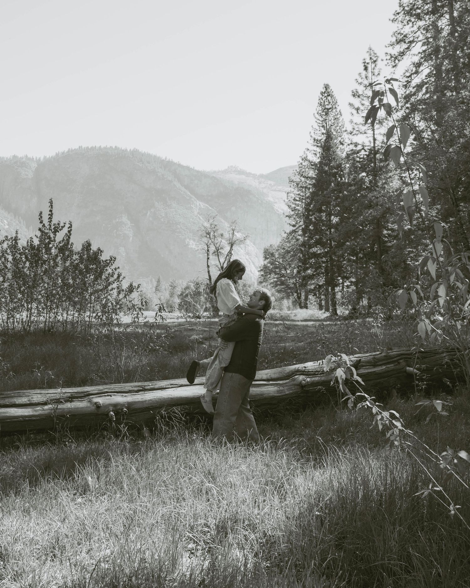 Black and white photo of couple embracing in rustic mountain forest meadow with fallen logs.