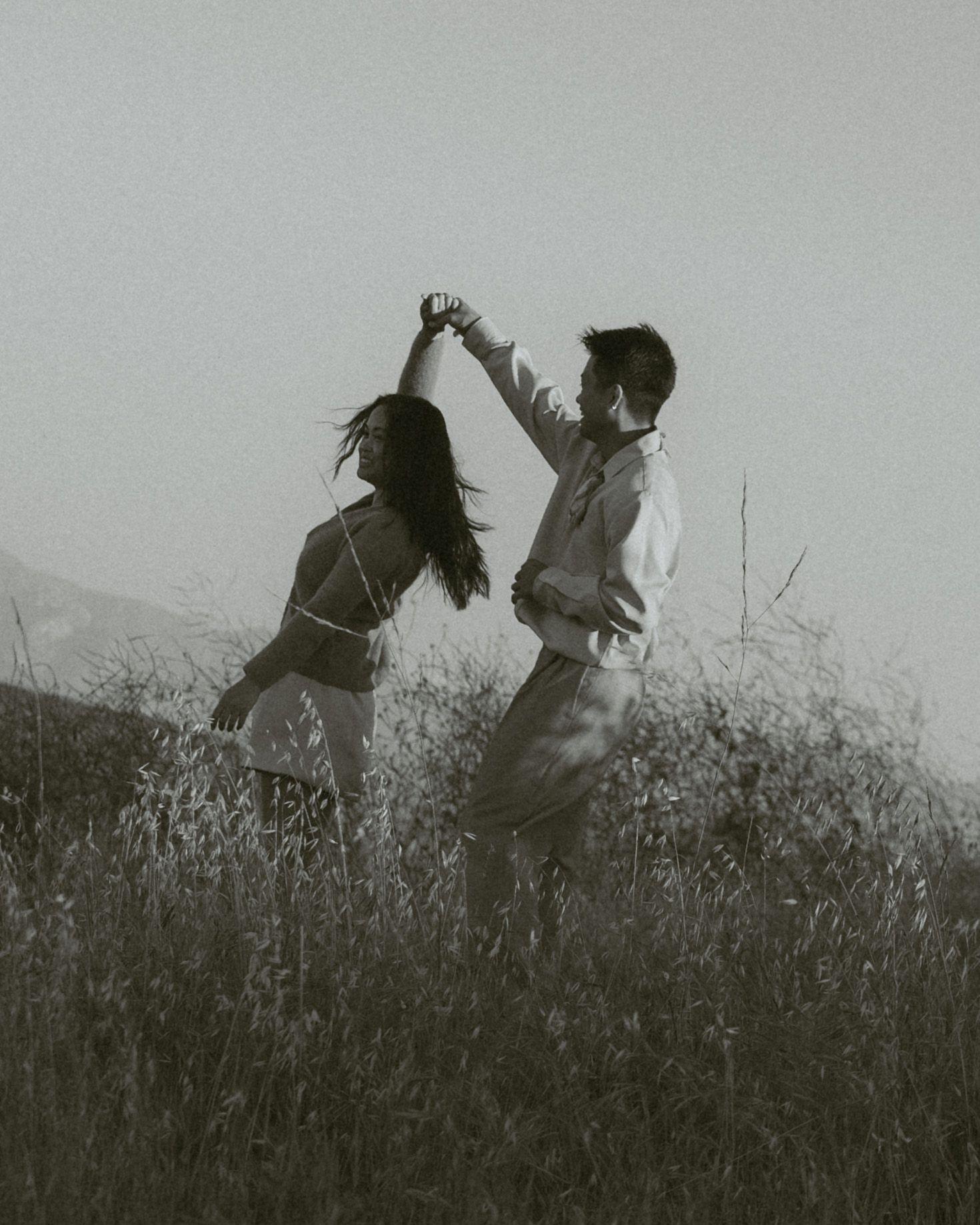 Black and white photo of a couple dancing romantically in a field, holding hands raised against a misty sky.