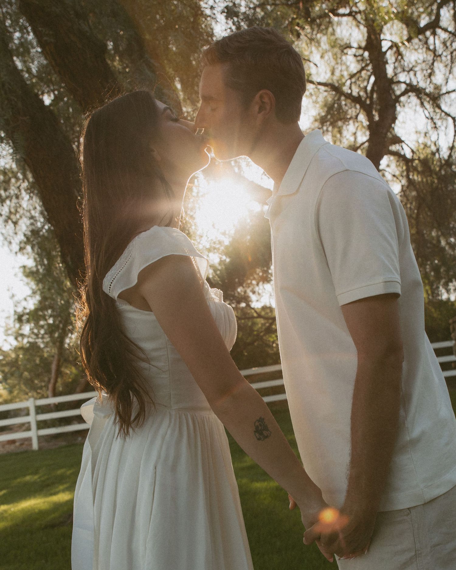 Couple kissing at golden hour sunset, woman in white dress and man in white polo, holding hands outdoors near trees.