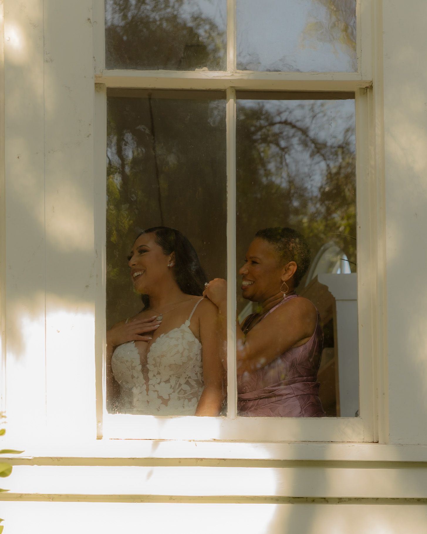 Bride in lace wedding gown smiling with bridesmaid in lavender dress, seen through a sunlit white window frame.