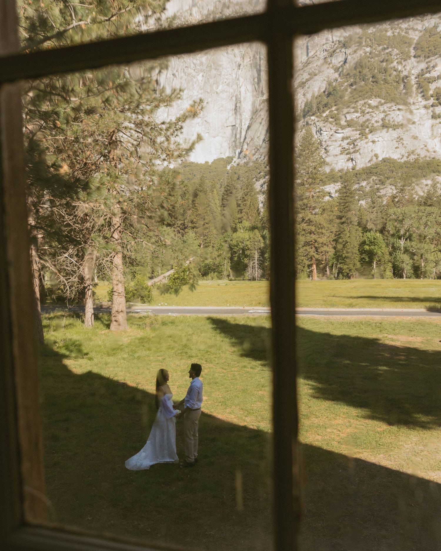 Couple in wedding attire holding hands in Yosemite meadow viewed through rustic window frame with granite cliffs beyond.