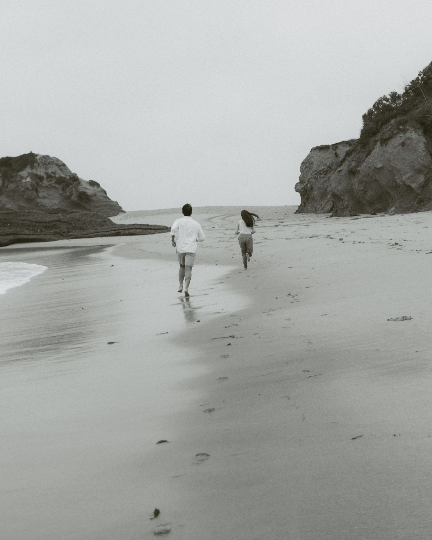 A couple runs hand-in-hand along a misty beach with rocky cliffs in a black and white photo.