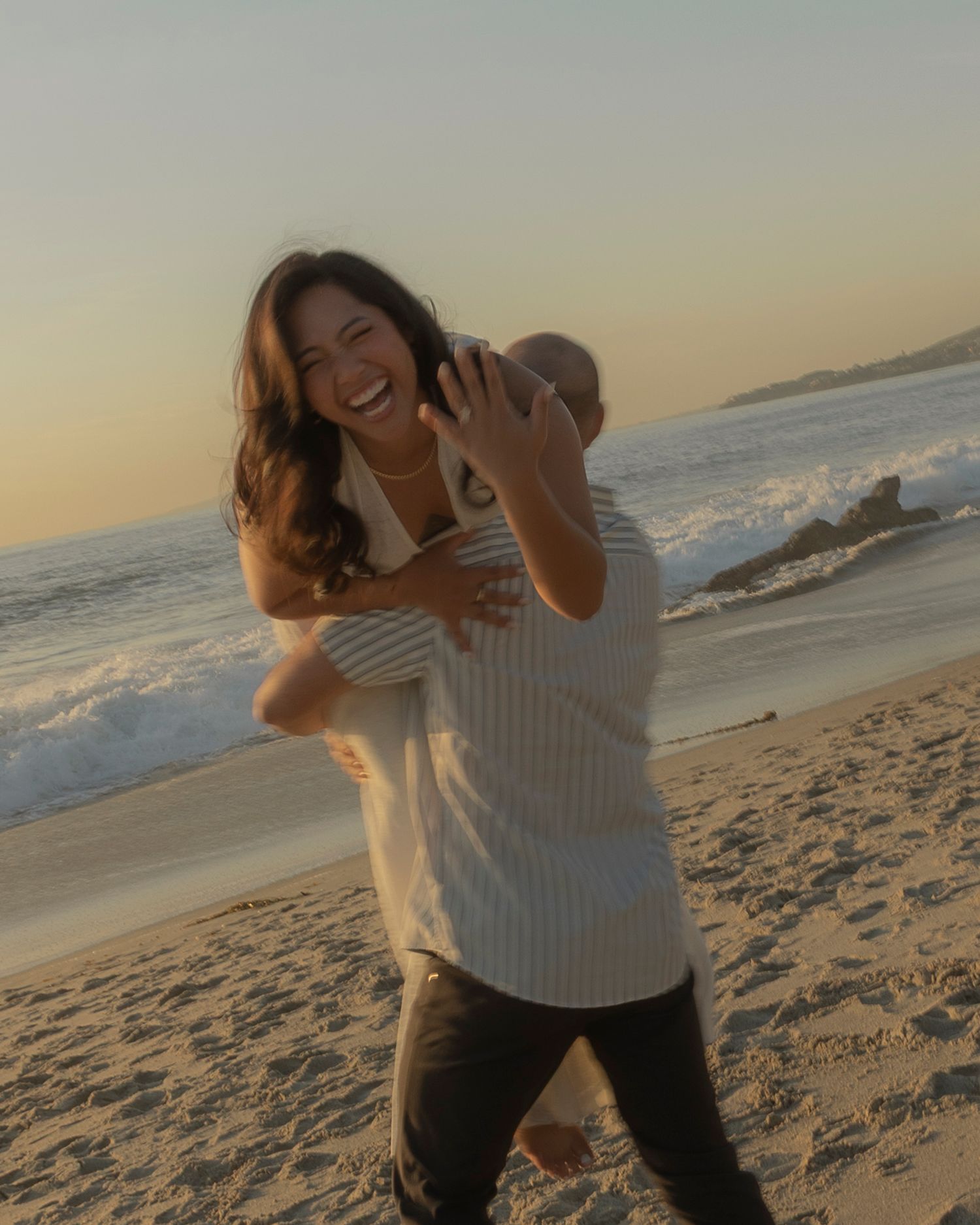 A person in a white sweater laughs and plays on a sandy beach at sunset with ocean waves in the background.